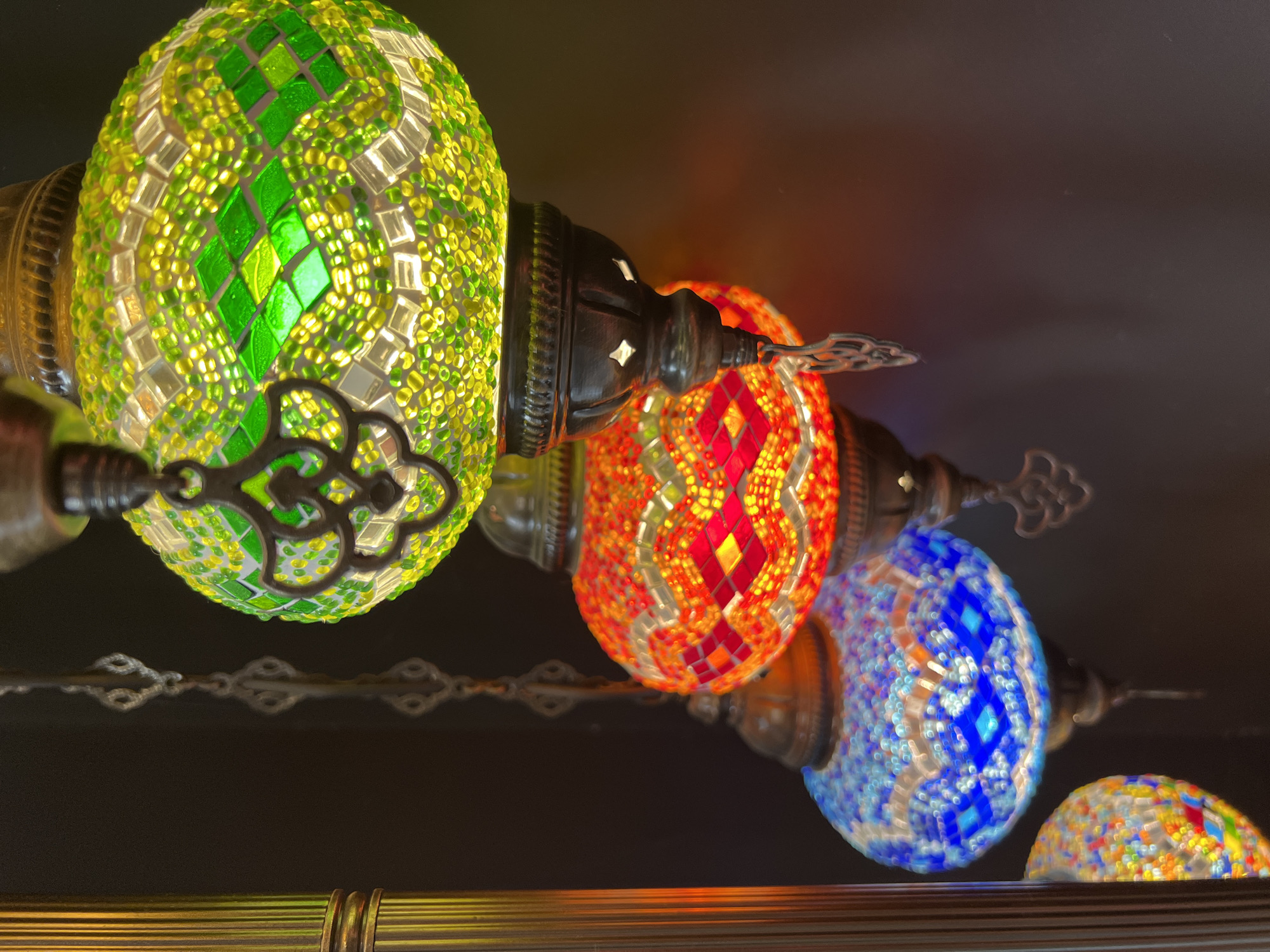 This media file features a close-up, low-angle shot of several decorative mosaic glass lamps. The lamps are oval-shaped and illuminated from within, casting a warm glow. From left to right, the visible lamps are primarily green, orange/red, and blue, each with intricate mosaic patterns of small glass pieces. The green lamp showcases diamond shapes and a checkerboard-like pattern of lighter and darker green and clear glass. The orange/red lamp displays a diamond pattern with red and orange glass. The blue lamp features a geometric pattern with various shades of blue and clear glass.
The lamps are suspended by dark metal fixtures and chains, suggesting they are part of a chandelier or ceiling light arrangement. The background is dark, with some ambient light reflecting off a reflective surface at the bottom of the frame, which appears to be a wooden or metallic shelf. The lighting suggests an indoor setting, possibly a shop or a decorative space, with the illumination coming from the lamps themselves and possibly overhead ambient light. The overall impression is one of warmth, color, and intricate craftsmanship. No people, text, or specific environmental conditions like weather or time of day are discernable. The location context is identified as Krasnodar, Russia.