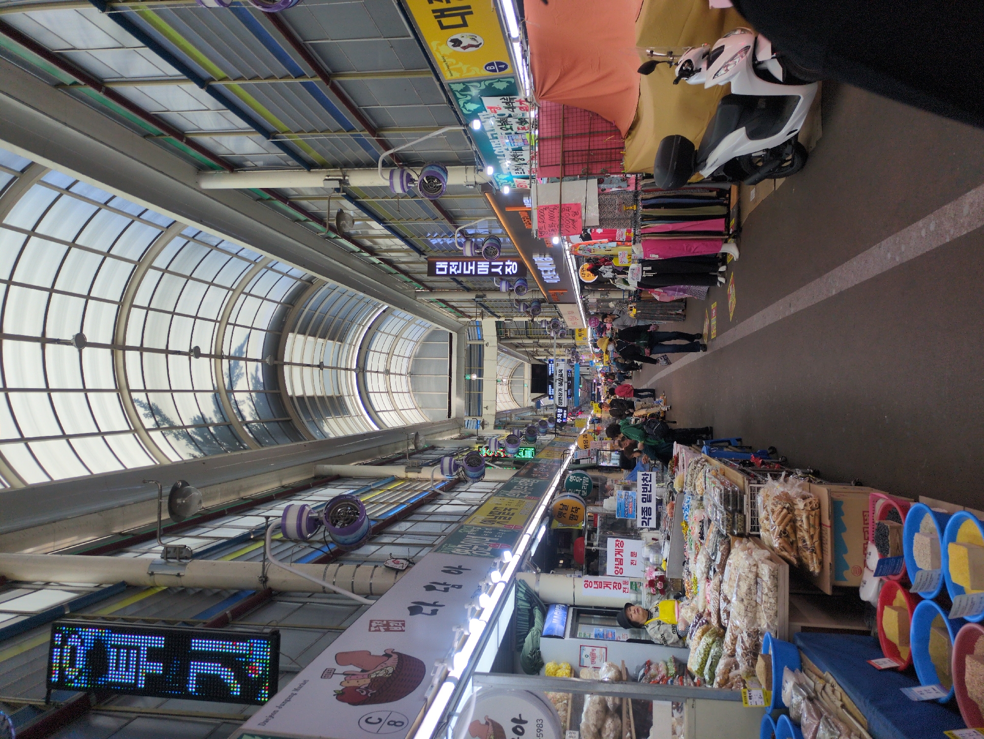 The image captures an indoor market scene in Daejeon, South Korea, specifically at Daejeon Jungang Market. The market is covered by a transparent arched roof structure. The aisle is lined with stalls on both sides. On the right side of the aisle, stalls display food items such as dried goods and prepared ingredients in colorful containers. People are seen walking through the aisle, some looking at the merchandise. On the left side, stalls display clothes and other goods. Signs and banners in Korean hang above the stalls. A parked scooter is visible on one side of the aisle. The environment is bustling with activity typical of a marketplace.