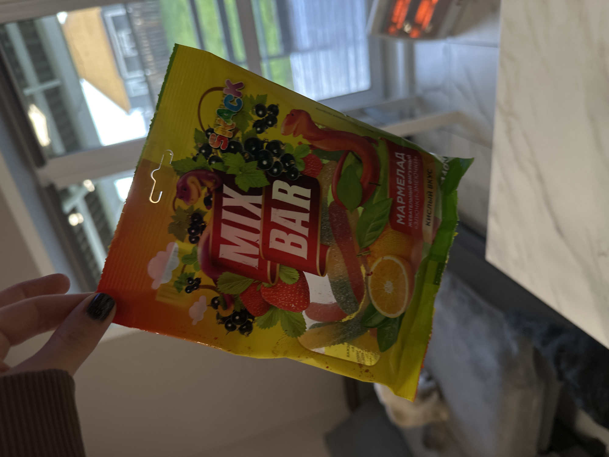 A hand with dark-painted fingernails holds a package of "Mix Bar" marmalade candies, featuring images of blackcurrants, strawberries, oranges, and long gummy worms. The packaging is predominantly yellow, orange, and green. The background includes a window with a view of trees, an overhead light fixture, and a gray countertop. The location is indeterminable; no reference to Kuda, Russia is visible in the image.