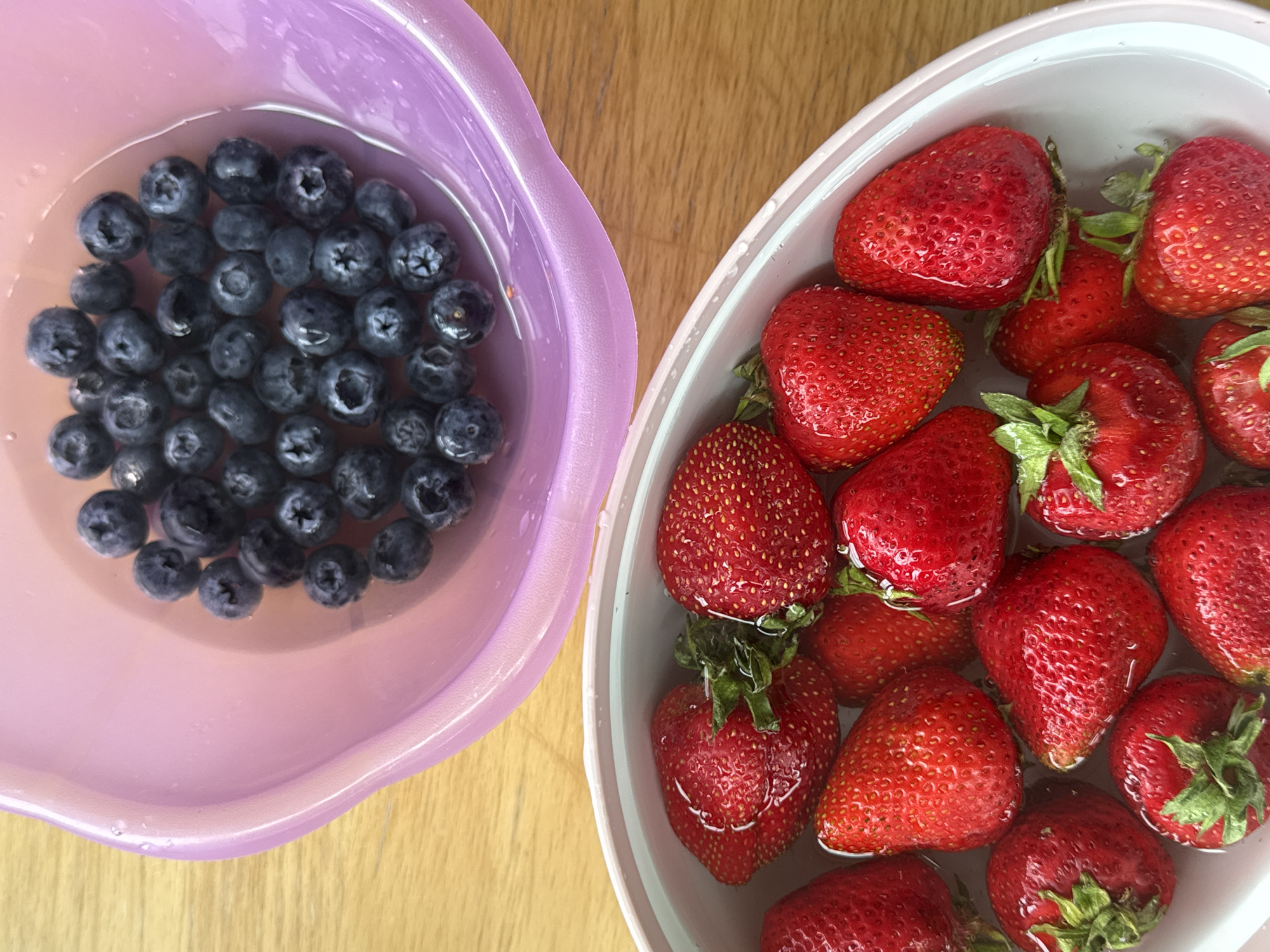 The image shows a top-down view of two bowls of fruit on a wooden surface. The upper bowl, which is lavender in color, contains numerous blueberries submerged in water. The lower bowl, which is light pink with a white interior, is filled with ripe strawberries, also in water. The lighting suggests an indoor setting, possibly a kitchen. There are no people or any indications of a specific city or location.