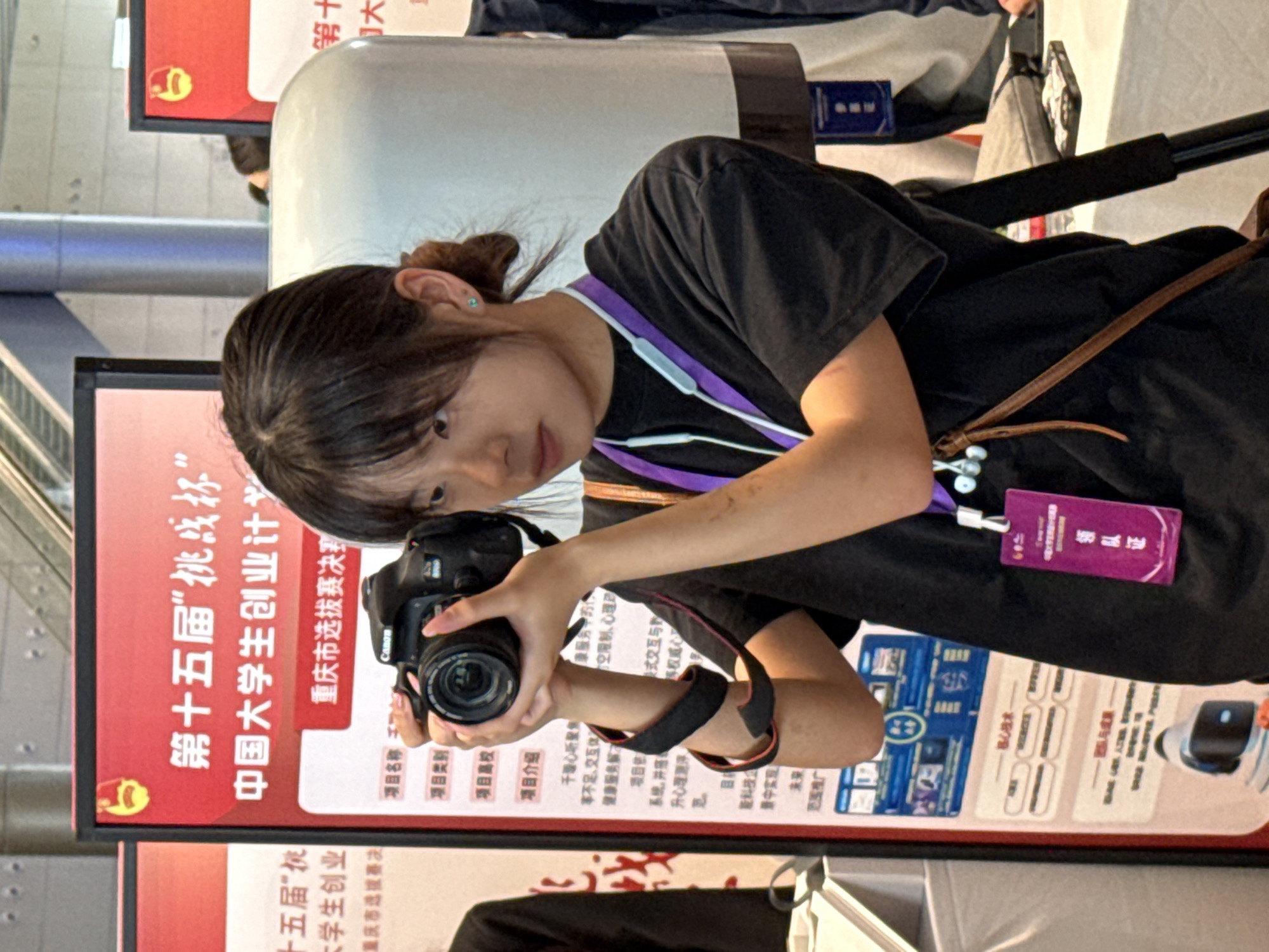 A young woman with dark hair pulled into a bun, wearing a black t-shirt and a purple lanyard, holds a black Canon DSLR camera up to her face, appearing to take a photo. She has a small turquoise earring in her left ear. She wears a brown crossbody bag with thin straps. A purple badge with Chinese characters is attached to her shirt.
The woman is standing in front of a large red display board with Chinese text. The text is difficult to read in full, but includes "第十五届”挑战杯”中国大学生创业计划" which translates to "The 15th 'Challenge Cup' China University Student Entrepreneurship Plan." Another sign to the left appears to be a continuation of this title.
Behind the woman, to her right, is a large, white, cylindrical object with a dark base. The environment suggests an indoor event, possibly an exhibition or competition, given the display boards and the woman's activity. The lighting is bright and even. The background shows blurry elements that could be other attendees or exhibition booths.
The specific location is identified as Chongqing High-tech Zone, China based on the event title referencing a competition relevant to the region.
