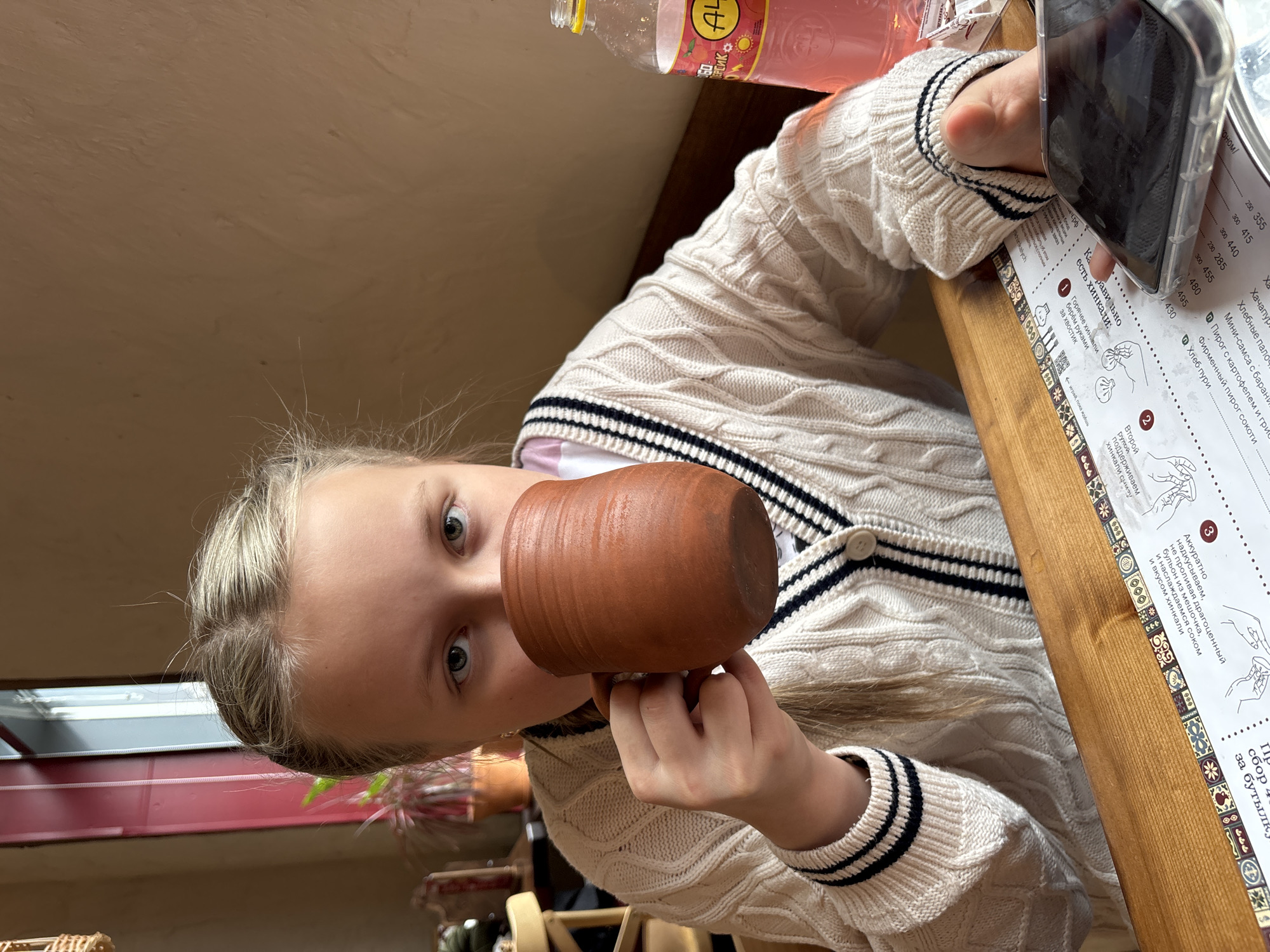 A young, fair-skinned person with blond hair is holding a brown clay mug and drinking from it, looking directly at the camera. The person is wearing a white cable-knit cardigan with dark blue and white striped trim. To the person's right is a menu card with text written in Russian, next to which is a smartphone. In the upper right corner of the frame, there is a bottle of juice. The image was taken in a restaurant/cafe in Izhevsk, Russia.