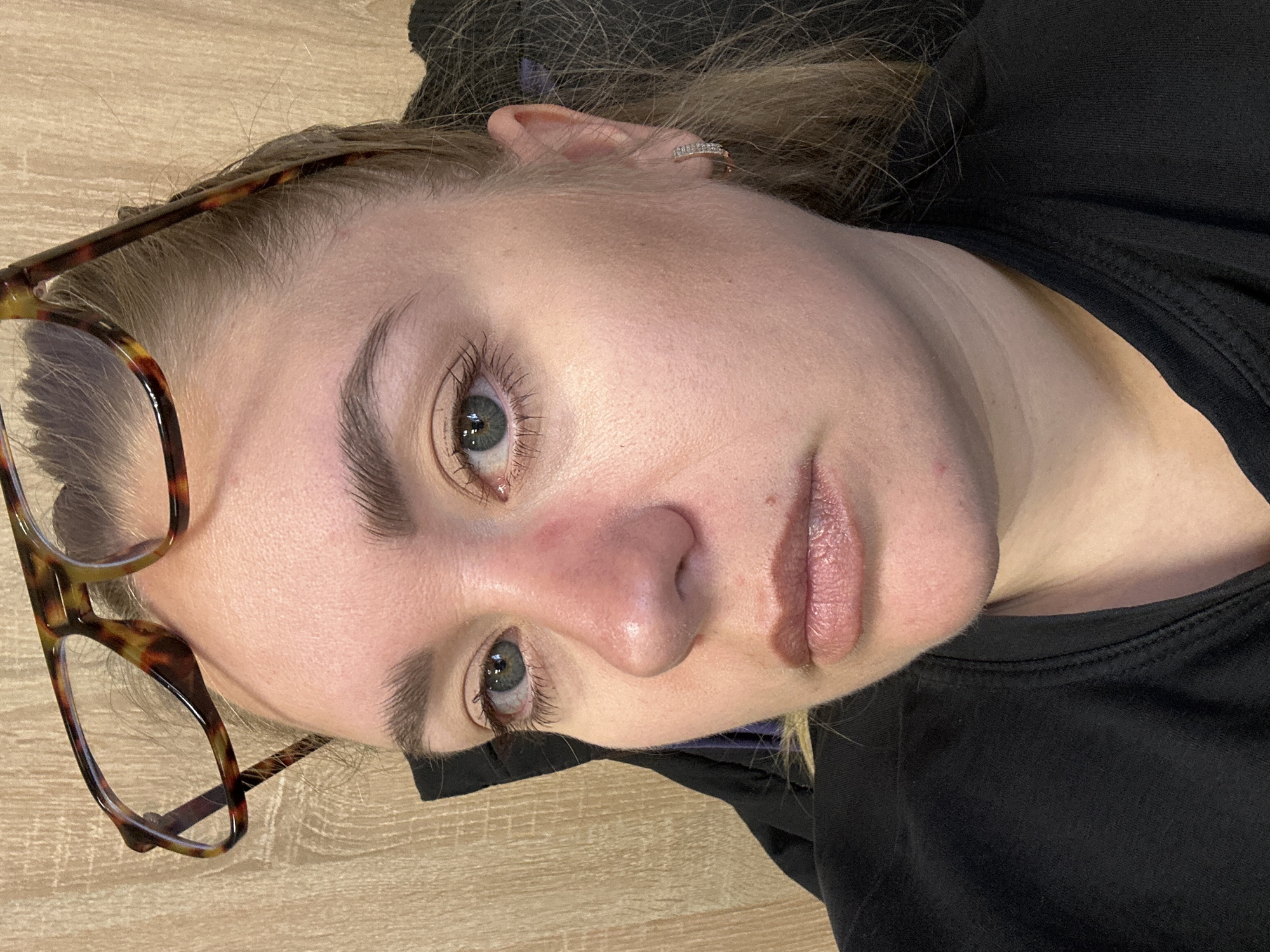 A close-up, eye-level portrait shot shows a fair-skinned woman with light-colored hair, possibly blonde or light brown. She is wearing glasses with a tortoise-shell pattern frame. Her eyes are a grayish-blue color. She has a black shirt or jacket on, suggesting cool weather. The background appears to be a light-colored wooden surface. No specific landmarks or details indicate the location as Ural'skoe, Russia.