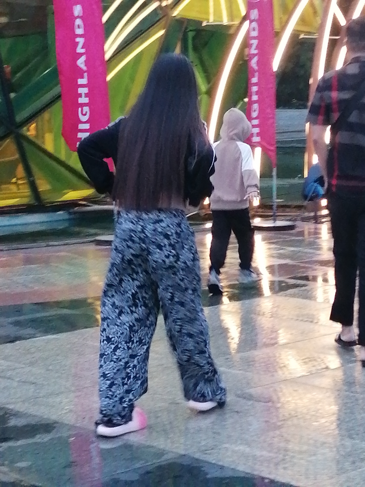 In Da Lat, Vietnam, a young woman with long dark hair stands facing away from the camera. She wears a black long-sleeved top and loose, patterned pants with a blue and white floral design. Her left foot is shod in a bright pink slip-on shoe, while her right foot is not fully visible. Behind her, a child in a light brown hooded jacket and dark pants walks away. To the far right, a person wearing a black and red plaid shirt and black pants is also visible. They stand on a wet, reflective surface, likely a plaza or walkway, illuminated by artificial lights from overhead structures, which appear to be part of a modern building. Two vertical pink banners with white lettering, one reading "HIGHLANDS," are positioned behind the central figures. The overall scene suggests an outdoor urban environment during or after rain.