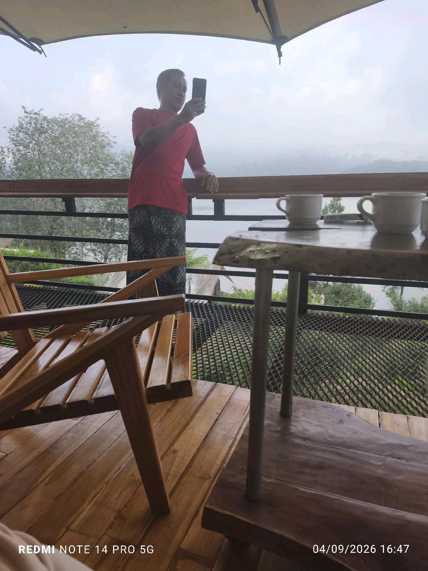 An elderly man in a red t-shirt and patterned pants stands on a wooden balcony, holding a smartphone and appearing to take a photo or video. In the foreground, a wooden chair is positioned on the balcony floor. A rustic wooden table with three white coffee cups sits on the balcony railing, overlooking a misty landscape of rolling hills and trees. The scene is dated 04/09/2026 16:47 and displays "REDMI NOTE 14 PRO 5G" at the bottom. The environment suggests a serene, possibly mountainous or hilly region.