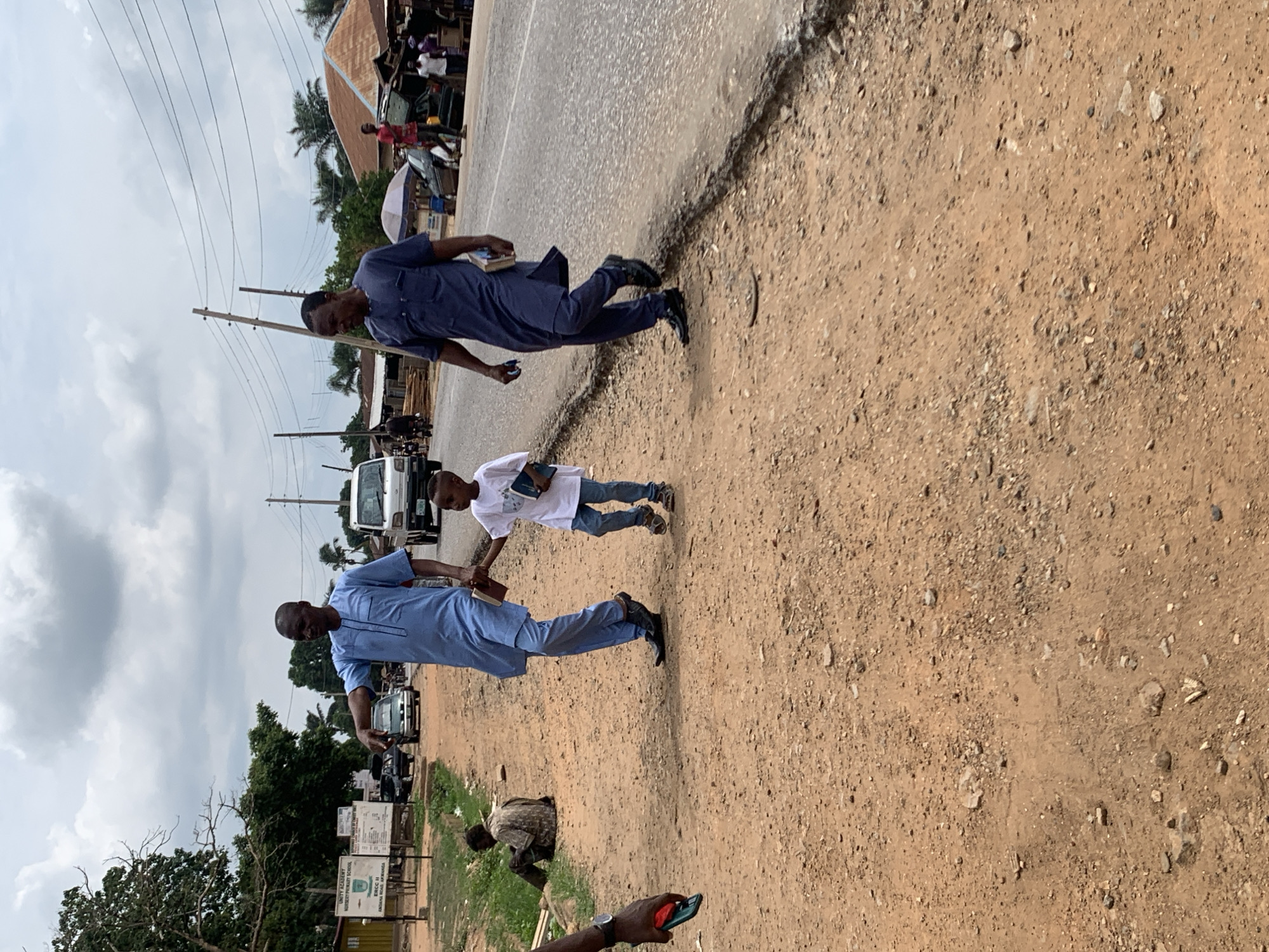 The image shows a street scene in Akwanga, Nigeria. Two men, one wearing a light blue traditional outfit and the other a dark blue one, walk along the side of the road, holding a child's hand between them. The child wears a white t-shirt and jeans. Both men and the child are holding what appear to be books or small notebooks. The road is paved, and the shoulder is unpaved dirt. In the background, there are parked cars, utility poles, and buildings with people visible. A sign reads "UNITY ACADEMY MOSKEY PRIMARY SCHOOL". Another man is near the ground near the sign. Another person is holding a cell phone in the foreground. The sky is overcast.