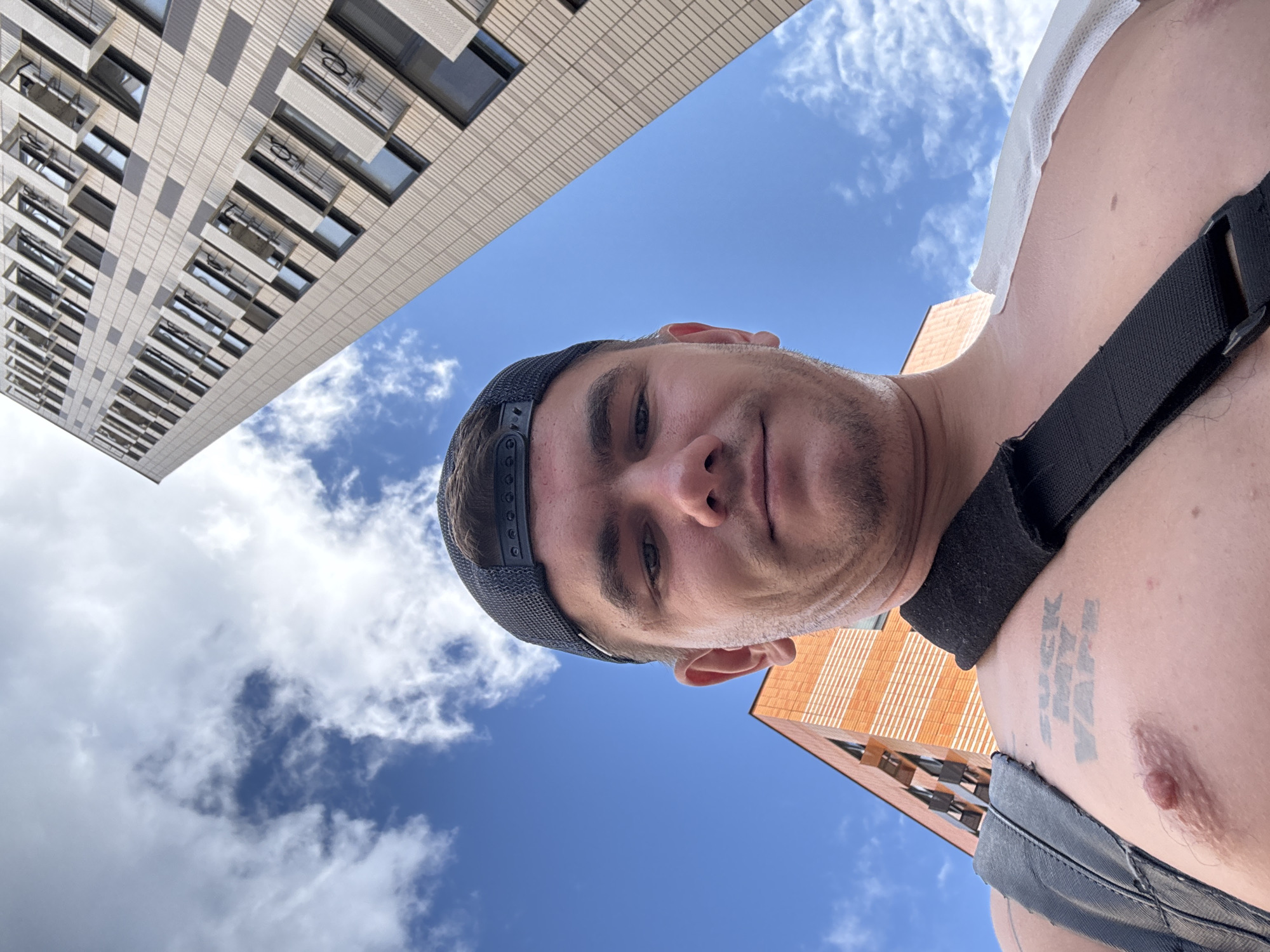 This is a selfie taken outdoors on a bright, cloudy day. A young man wearing a black baseball cap and no shirt is the primary subject. He has a tattoo on his chest that reads "FUCK MY VAPE." A black strap crosses his chest, and there is a white bandage on his right shoulder. In the background, two modern apartment buildings are visible against a blue sky with scattered white clouds. The architecture of the buildings suggests an urban environment, potentially in Moscow, Russia, given the context provided. One building is predominantly orange brick with white accents, while the other is a taller, beige structure with many windows. The perspective is from below, looking up at the man and the buildings.