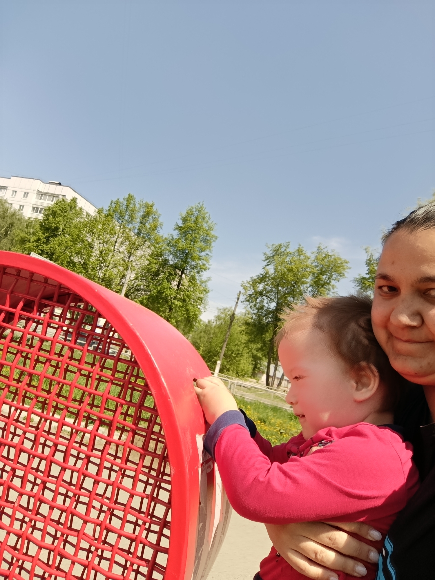 A medium shot shows a woman holding a young child at an outdoor playground on a sunny day, likely in Chaikovsky, Russia. The child, wearing a red long-sleeve shirt with blue trim, is reaching toward a large, red, spherical climbing structure made of a grid-like netting. The woman is smiling gently at the camera. In the background are trees, a white apartment building, and a clear blue sky. The ground appears to be grassy.