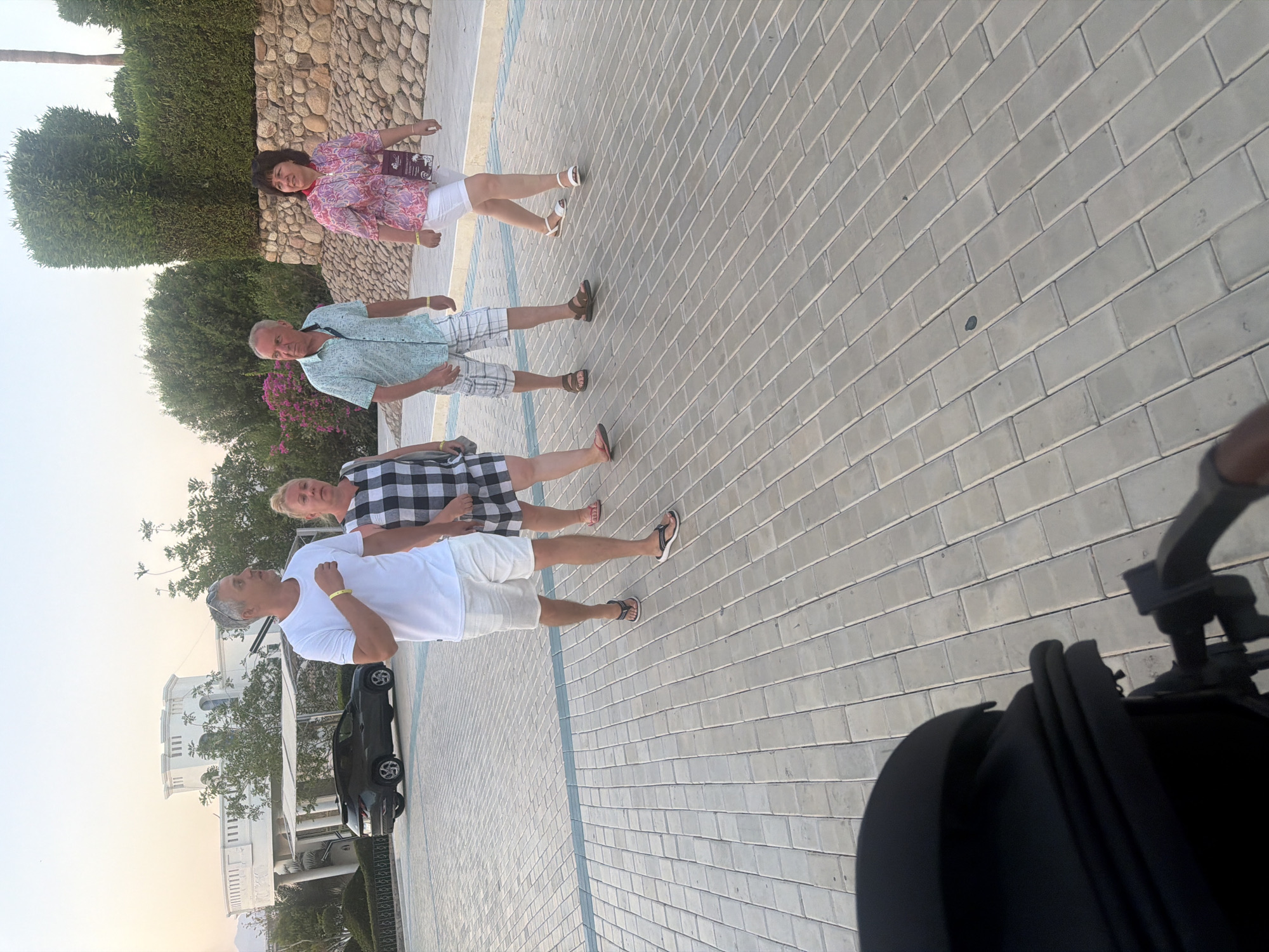 The image shows a paved walkway in Sharm El Sheikh, Egypt, with four individuals walking away from the camera. The ground is covered in light-colored rectangular pavers. To the left, a white SUV is parked, and a modern white building with decorative turrets is visible in the background, surrounded by green foliage. Further to the right, a stone wall built with large, rounded rocks is visible, with manicured green hedges atop it.
The individuals are walking in a line. The person furthest to the left is a man wearing a white t-shirt and white shorts, with black flip-flops. Next to him is a woman in a black and white checkered dress. To their right is a man in a light blue patterned short-sleeved shirt and checkered shorts, wearing brown sandals. The woman furthest to the right is wearing a patterned pink and white top and white shorts with white sandals.
The sky is a pale orange, suggesting either sunrise or sunset. In the foreground, a portion of a black stroller is visible, indicating that the photo was likely taken from the perspective of someone pushing a baby carriage.