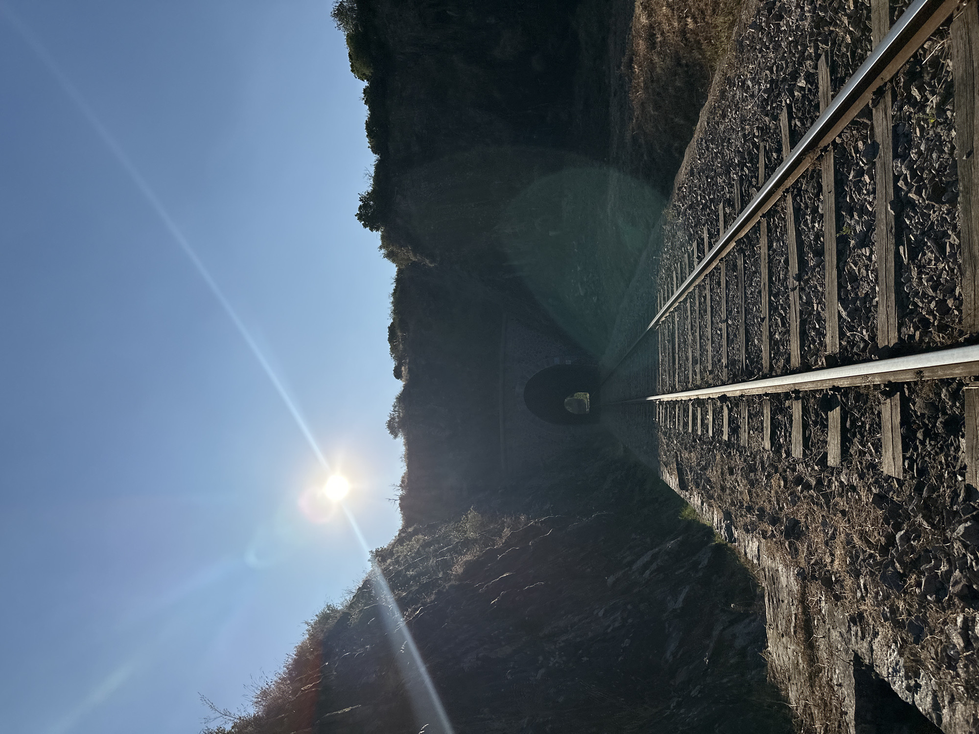 The image depicts a set of railroad tracks leading towards a stone tunnel, nestled between rocky, vegetated embankments under a clear blue sky with the sun shining brightly, causing lens flare. The sun is positioned in the upper left quadrant of the image, casting long shadows. The railroad tracks are made of metal rails supported by wooden sleepers, laid on a bed of ballast. The tunnel entrance is dark and arched, with a faint light visible at the end, suggesting it leads to an open area or another scene beyond. The environment appears to be rural or semi-rural, with dry vegetation and rocks visible on the slopes. The scene suggests an abandoned or disused railway line, given the natural overgrowth and the weathered appearance of the stone structure. The location is identified as Générargues, France. There are no people or animals visible in the image.