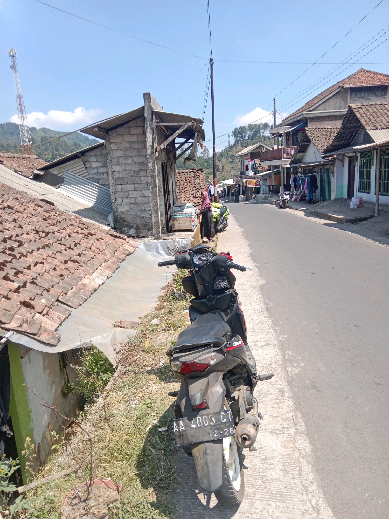 The image depicts a street scene in Adipuro, Indonesia, likely a rural or semi-rural area, with a paved road running through it. In the foreground, a dark-colored motorcycle with a license plate reading "AA 4003 CT" is parked on the side of the road. The road is bordered by a sidewalk or pathway made of concrete. On the left side of the road, there are several buildings with tiled roofs, some appearing to be houses or small shops. One building is constructed with cinder blocks and has a corrugated metal roof. Vegetation, including grass and some small trees, grows along the roadside and in front of the buildings.
Across the street, more buildings are visible, some of which appear to be shops displaying clothing on racks outside. A few other motorcycles are parked along the sidewalk in front of these establishments. In the background, hills covered with trees rise under a clear blue sky with a few white clouds. A tall transmission tower with red and white markings is visible on one of the hills. The overall impression is of a sunny day in a developing urban or village setting.