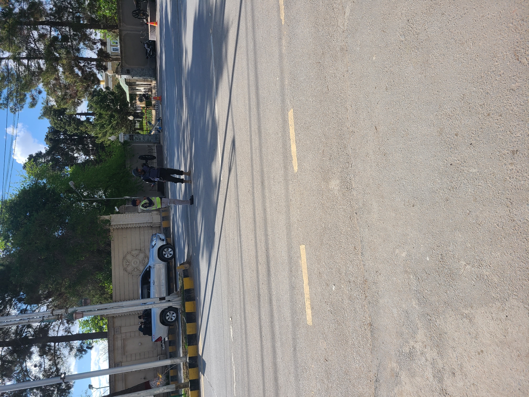 The image shows a street in Quetta City Tehsil, Pakistan. Two men are standing near the roadside. One is wearing a dark outfit with a helmet, and the other is wearing a high-visibility vest. A silver pickup truck is parked nearby along a concrete wall, which has some decorative elements. The road has yellow lane markings. Tall trees cast shadows across the road. Power poles with wires are visible in the foreground. Buildings and vegetation are present in the background.