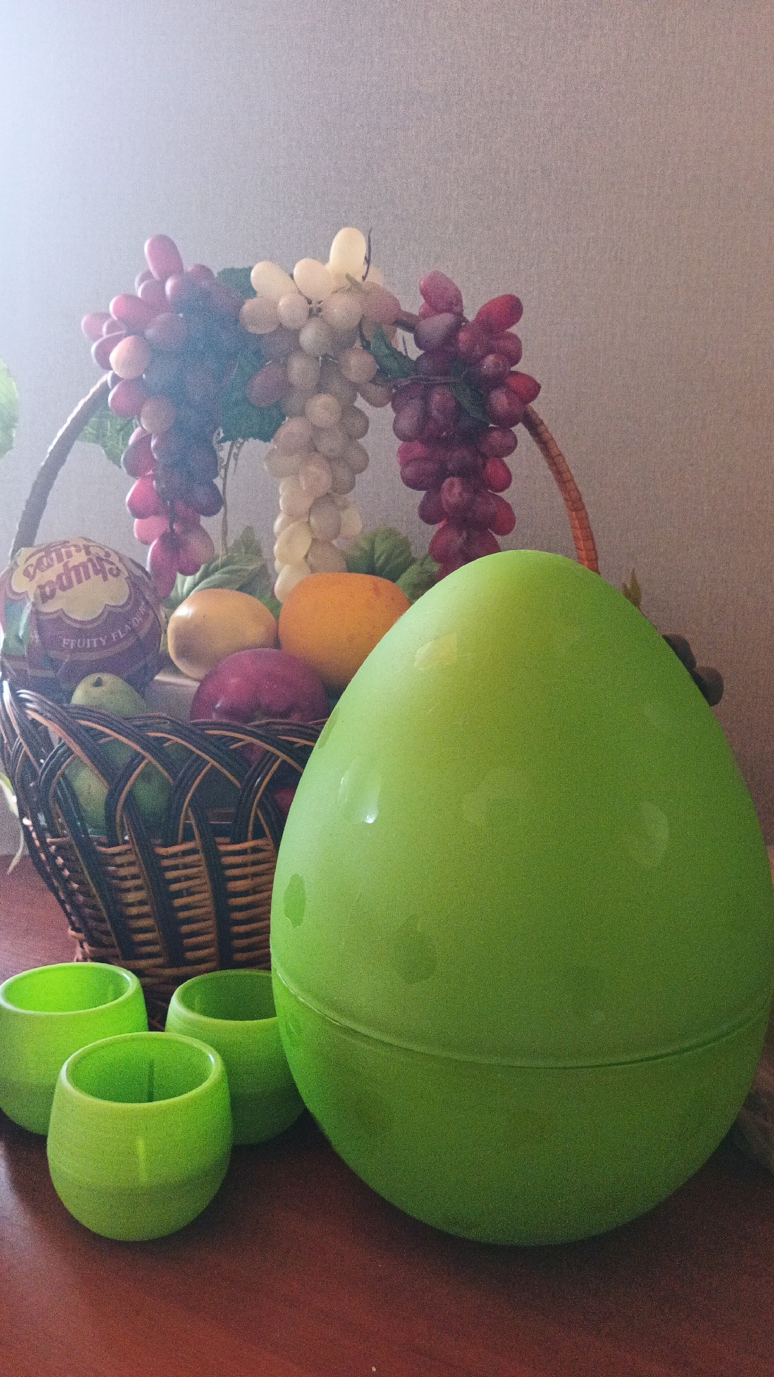 A still life arrangement features a wicker basket overflowing with artificial grapes in shades of red and white, along with plastic fruits including an orange and a plum. To the right of the basket, a large, bright green plastic egg rests on a dark wooden surface. In the foreground, three smaller, lime green plastic cups are arranged to the left of the egg. The background is a plain, textured gray wall. The lighting suggests an indoor setting, possibly a table or shelf. The image does not depict any elements that can be used to determine its location in Kryvyi Rih, Ukraine, or any other specific geographical location.