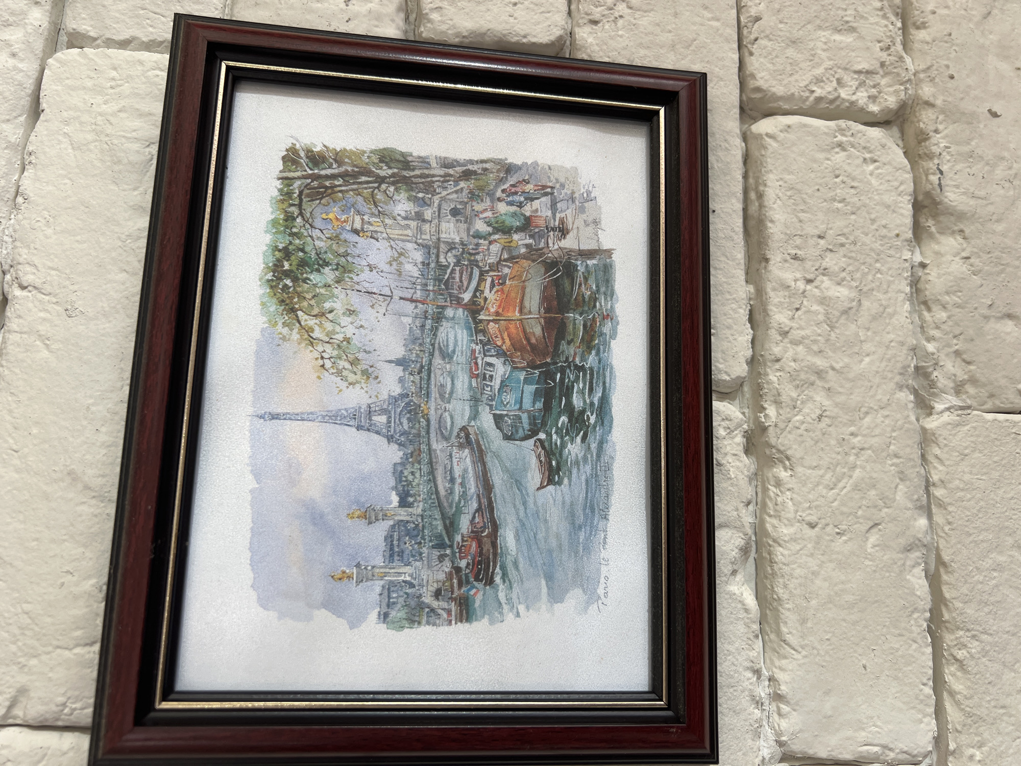 A framed watercolor painting is centrally positioned, mounted on a white-painted brick wall. The painting depicts a Parisian cityscape focusing on the Seine River, specifically featuring the Pont Alexandre III and the Eiffel Tower in the background. The bridge is characterized by its distinct golden statues.
On the river, a reddish-brown barge with a French flag on its stern is visible, alongside a small wooden rowboat. Two larger boats, one blue and one reddish-orange, are docked on the right bank. The right bank features a tree-lined promenade where several small, indistinct figures are walking. The sky above is rendered with light blue and grey watercolor washes. In the bottom left corner of the painting, the title "Paris, le Pont Alexandre III" is inscribed in script.
The painting is enclosed in a dark reddish-brown wooden frame, which has a thinner black inner border accented by a thin gold line. The background wall consists of irregularly shaped and textured white-painted bricks, with prominent mortar lines.