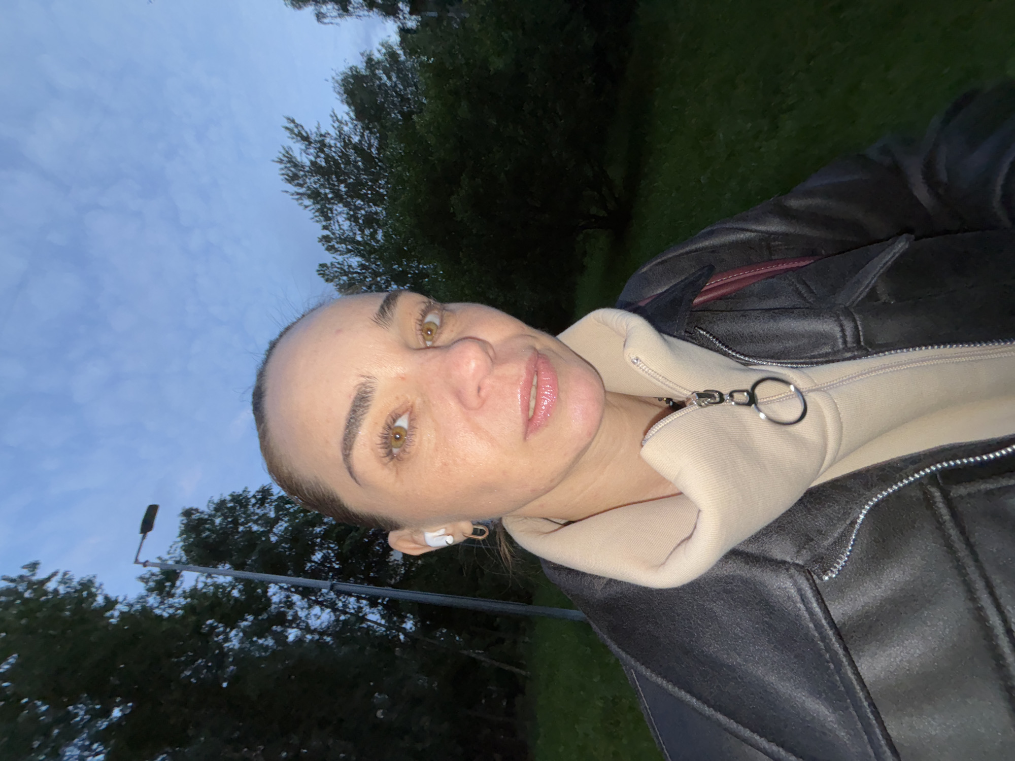 A close-up, selfie-style shot shows a woman in an outdoor environment with trees and a cloudy sky in the background. The woman has her hair pulled back and is wearing white earbuds and hoop earrings. She is dressed in a black leather jacket over a light-colored hooded sweatshirt with a silver zipper pull. The image appears to have been taken during twilight.