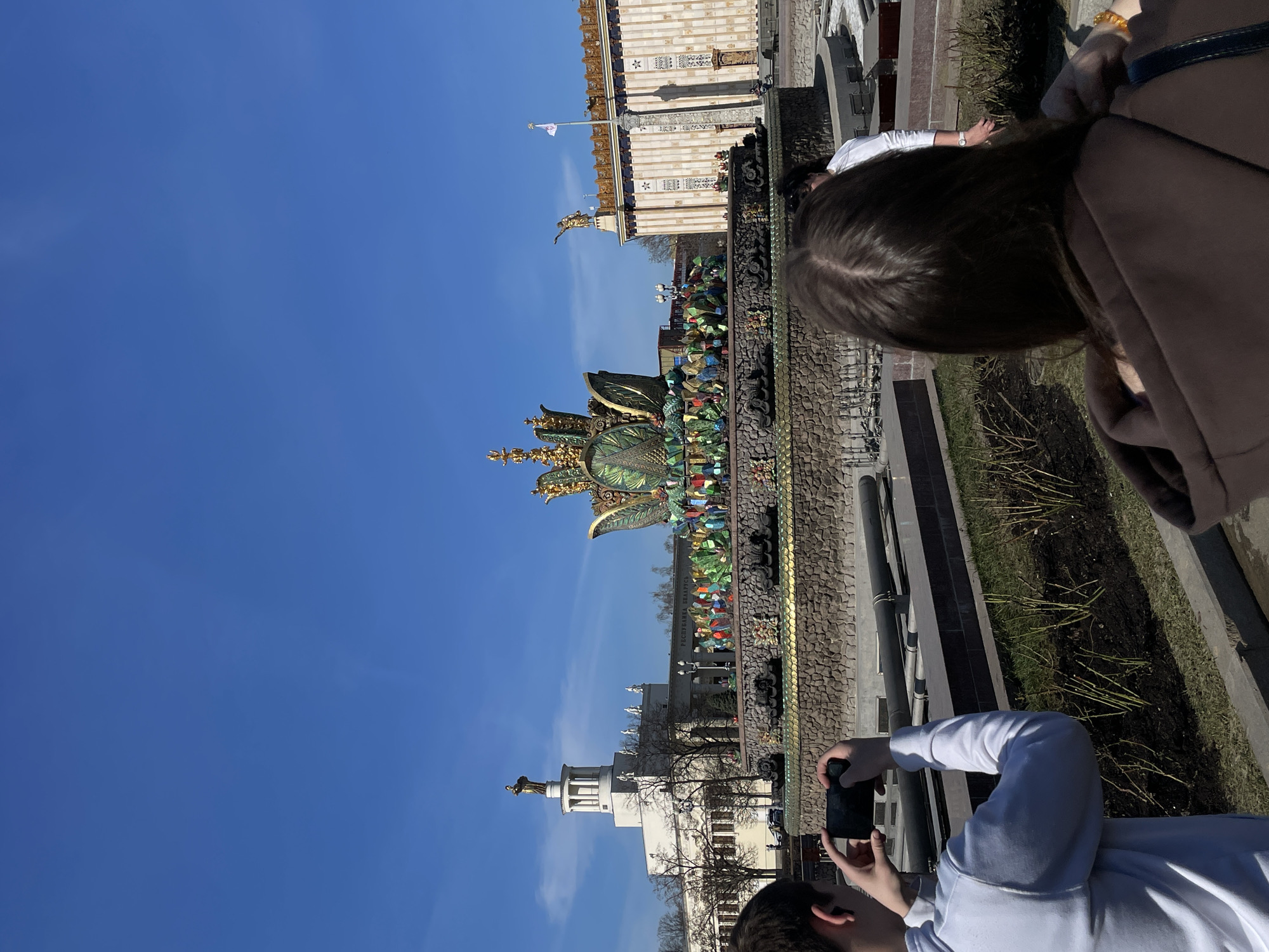 The image shows a tilted view of the People's Friendship Fountain at VDNH (Exhibition of Achievements of National Economy) in Moscow, Russia. The fountain is a prominent feature with its golden wheat stalks and various colorful mosaic elements. Buildings with statues on top can be seen in the background. Two people are visible in the foreground: one taking a photo with a smartphone and another standing next to them. The weather appears sunny, as indicated by the clear blue sky.