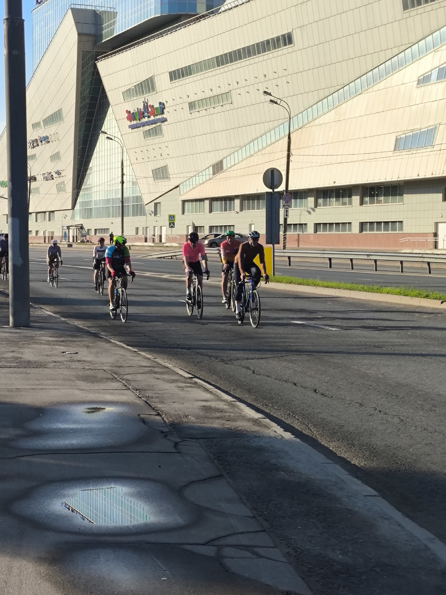 This image captures an urban scene featuring a group of cyclists on a wide road, set against the backdrop of a large, modern building.
**Subjects:** The primary subjects are a group of approximately seven to eight cyclists, all wearing helmets and athletic cycling attire, including brightly colored jerseys and shorts. They are riding road bicycles. In the background, a striking, multi-story building dominates the skyline. Other objects include streetlights, a traffic sign, and guardrails lining the roadside. The foreground features a concrete sidewalk with several puddles of water.
**Activity or event taking place:** The cyclists are actively riding their bicycles in a group, suggesting a recreational ride, training, or commute. They are spread across the width of the road, moving in the same direction.
**Notable details:** The long shadows cast by the cyclists and the building indicate that it is either early morning or late afternoon, likely morning given the bright, clear sky. The weather appears sunny and pleasant. The architectural style of the background building is distinctly modern, with angular lines, varying window shapes, and a facade that appears to be made of light-colored panels and extensive glass. One of the puddles in the foreground clearly reflects the building's geometric structure. The road is asphalt and seems to be a major urban thoroughfare.
**Visible text:** On the large building in the background, colorful, stylized text is visible, which appears to read "Jump City," suggesting it might be an entertainment complex or a commercial center.