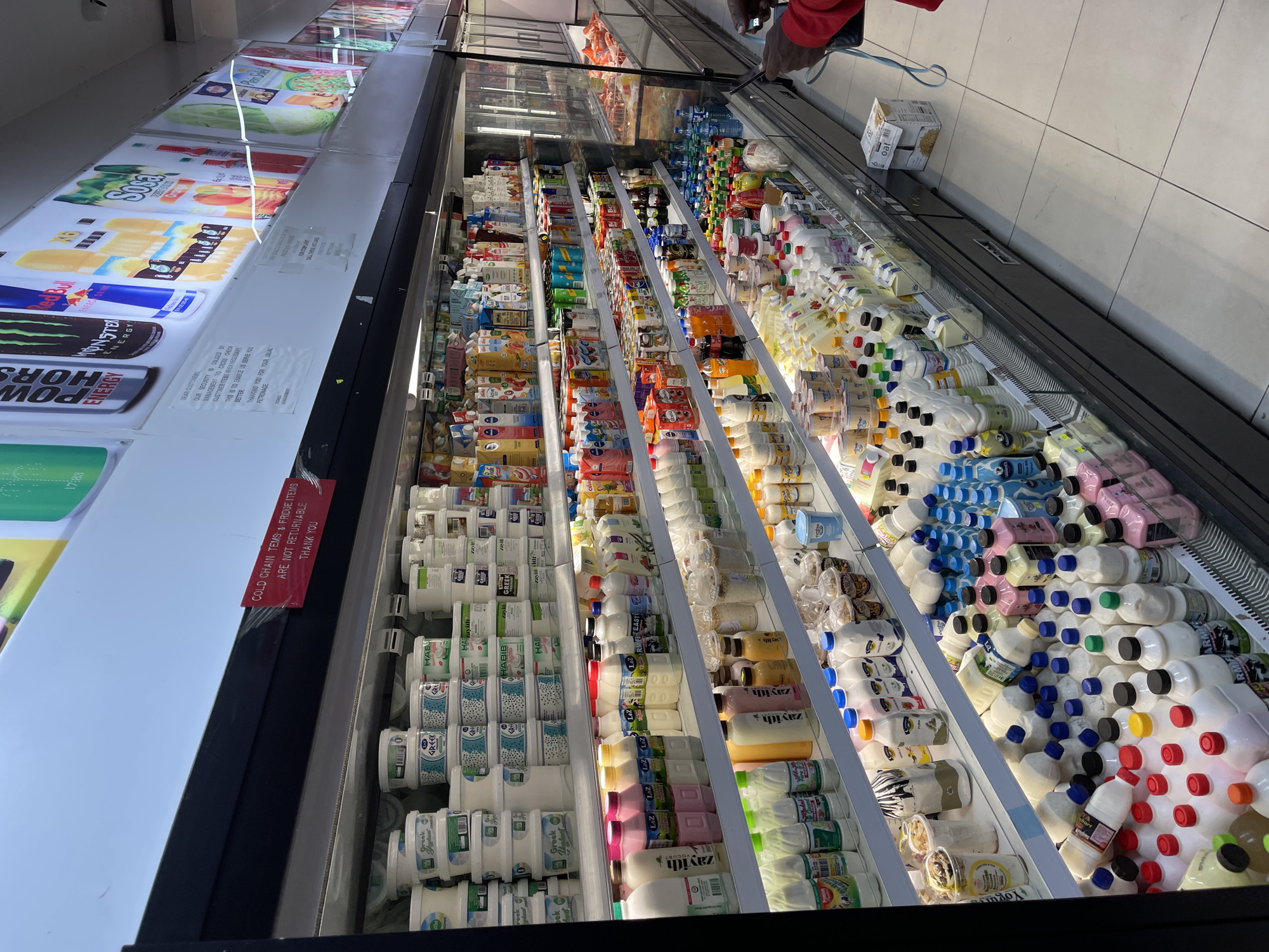 This is a photograph taken inside a well-stocked refrigerated display case in a grocery store located in Guzape, Nigeria. The display case is filled with a wide variety of dairy products, including milk, yogurt, and other beverages, packaged in various bottles, cartons, and tubs. The products are neatly arranged on multiple shelves, with a focus on beverages and yogurts at the bottom. The lighting is bright, illuminating the products and creating a clean, professional appearance. In the background, a person is visible, partially obscured by the display case, holding a smartphone. The overall environment suggests a typical retail setting where perishable goods are kept chilled for sale.