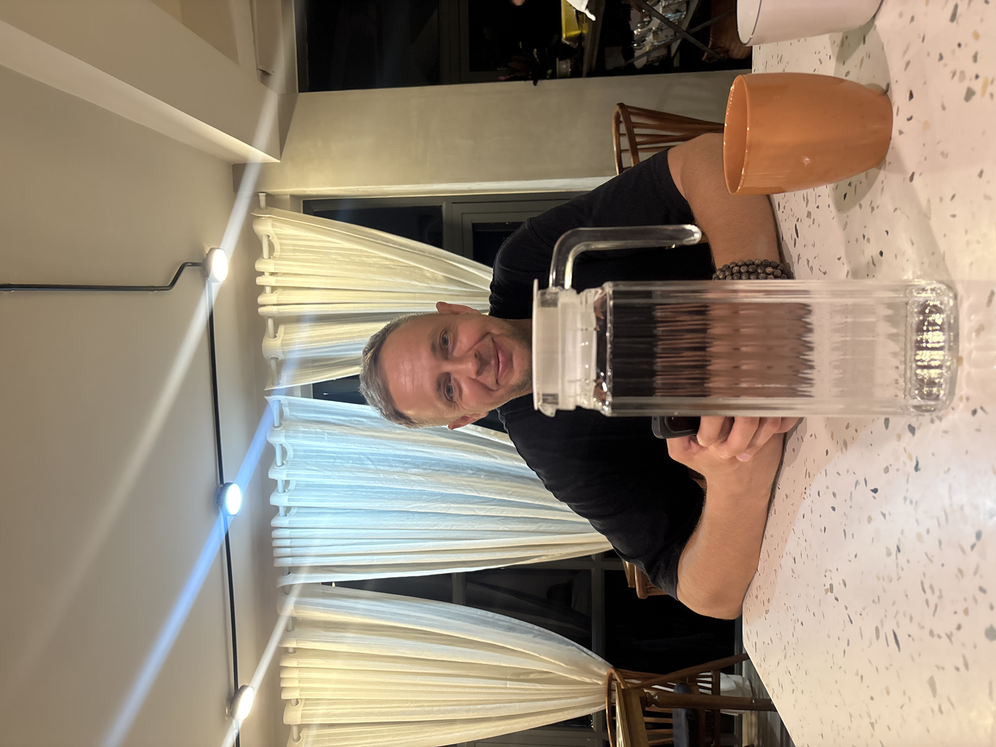 A man with short, graying hair and blue eyes is seated at a white speckled table, facing the camera with a slight smile. He is wearing a black short-sleeved shirt. His forearms rest on the table, and his hands are clasped in front of him, holding a dark-colored smartphone. On the table directly in front of him is a tall, clear glass pitcher filled with water, featuring a white lid and handle. To the right of the pitcher, an orange ceramic cup is visible, and partially cropped on the far right is a white cup. The man's left wrist wears a beaded bracelet.
In the background, a series of light-colored, possibly cream or off-white, pleated curtains are drawn, covering large windows. Through the gaps in the curtains and above them, dark window frames and the dark exterior are visible, suggesting it is nighttime or a dimly lit area outside. Wooden chairs with slatted backs are present in the midground, both to the left and right of the man. The ceiling above is light-colored and features several track lights. Two distinct light beams, one yellowish and one bluish, emanate from two of these circular light fixtures on a black track, projecting onto the ceiling. A structural beam or pillar is visible on the right side of the image, and further in the background to the right, some shelving or storage with various items can be vaguely discerned. The overall lighting suggests an indoor evening setting. Location: Unknown, Unknown.