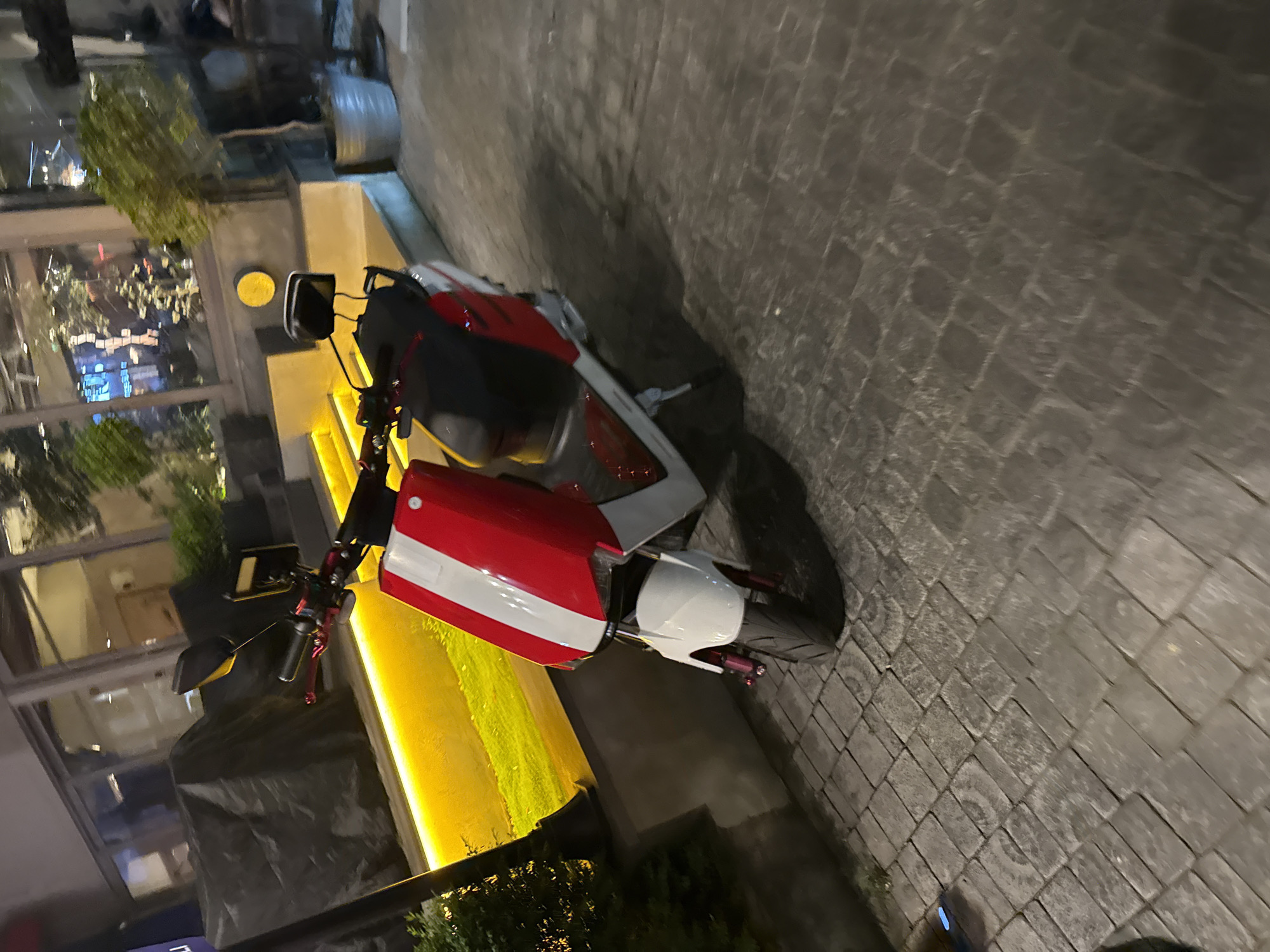 The image shows a red and white electric scooter parked on a brick-paved sidewalk at night. The sidewalk is bordered by a building with large glass windows reflecting interior lights and greenery, suggesting a restaurant or retail space. A yellow light source illuminates the scooter and the wall behind it. The photograph was taken in Yerevan, Armenia.