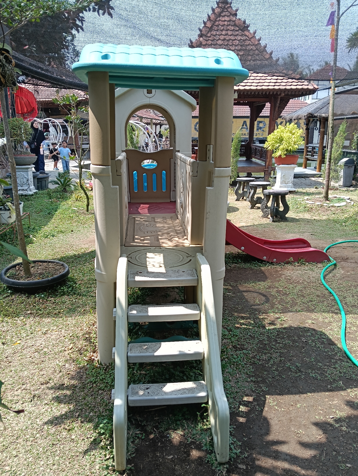 The image depicts a playground structure with a slide and stairs, set in an outdoor environment. The playground appears to be made of plastic and has a beige and blue color scheme. A red slide extends from the structure. The ground is covered with grass and dirt. In the background, there are some buildings with traditional Indonesian-style roofs and some trees. A green garden hose is visible on the right side of the image. Several people are present in the background, including a woman wearing a black hijab and two children. The scene is likely in a public area or a park.