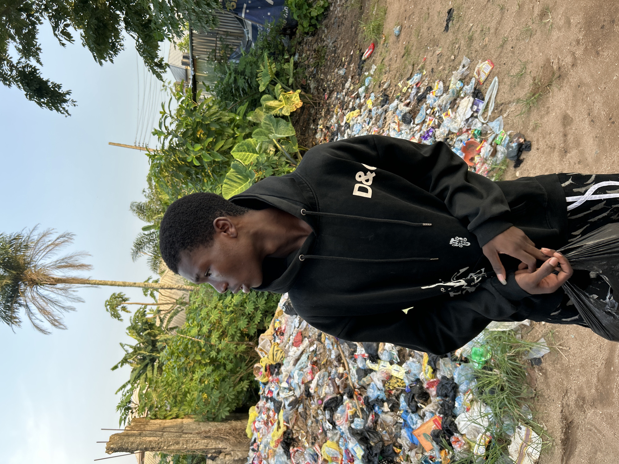 A young man in a black hoodie stands next to a large pile of mixed garbage in Ihiagwa, Nigeria. The garbage consists of plastic bags, food wrappers, and other discarded items. Behind the garbage are trees, plants, and a building. The ground is dirt and sand. The sky is light blue.