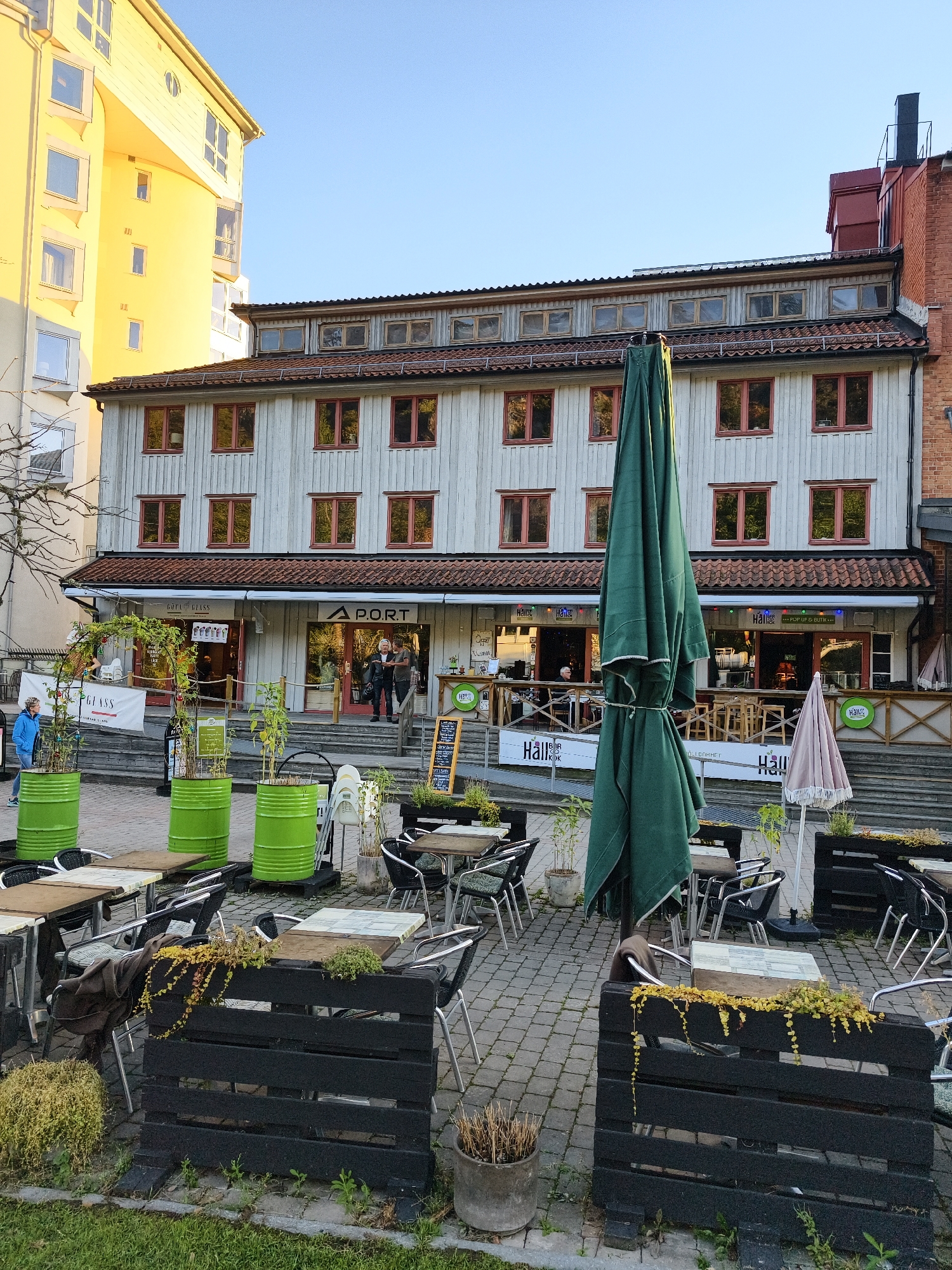 This is an eye-level, medium shot taken outdoors on a sunny day in Söderköping, Sweden. In the background, a light-colored, multi-story building with a mansard roof and many uniformly spaced windows with red frames dominates the scene. A bright yellow, modern building is partially visible to the left of the light-colored building. In the foreground, an outdoor seating area for a restaurant or cafe is set up on a cobblestone patio. There are several tables and chairs, some with umbrellas or large, folded green umbrellas. Decorative elements include large green cylindrical planters and black wooden planters with plants. A woman in a blue jacket walks past on the left side of the frame, and two men stand on the steps of the building in the center. Signs for "A.PORT" and "GULA GLASS" are visible on the building's facade, indicating businesses are located there. The overall impression is a vibrant street scene with outdoor dining.