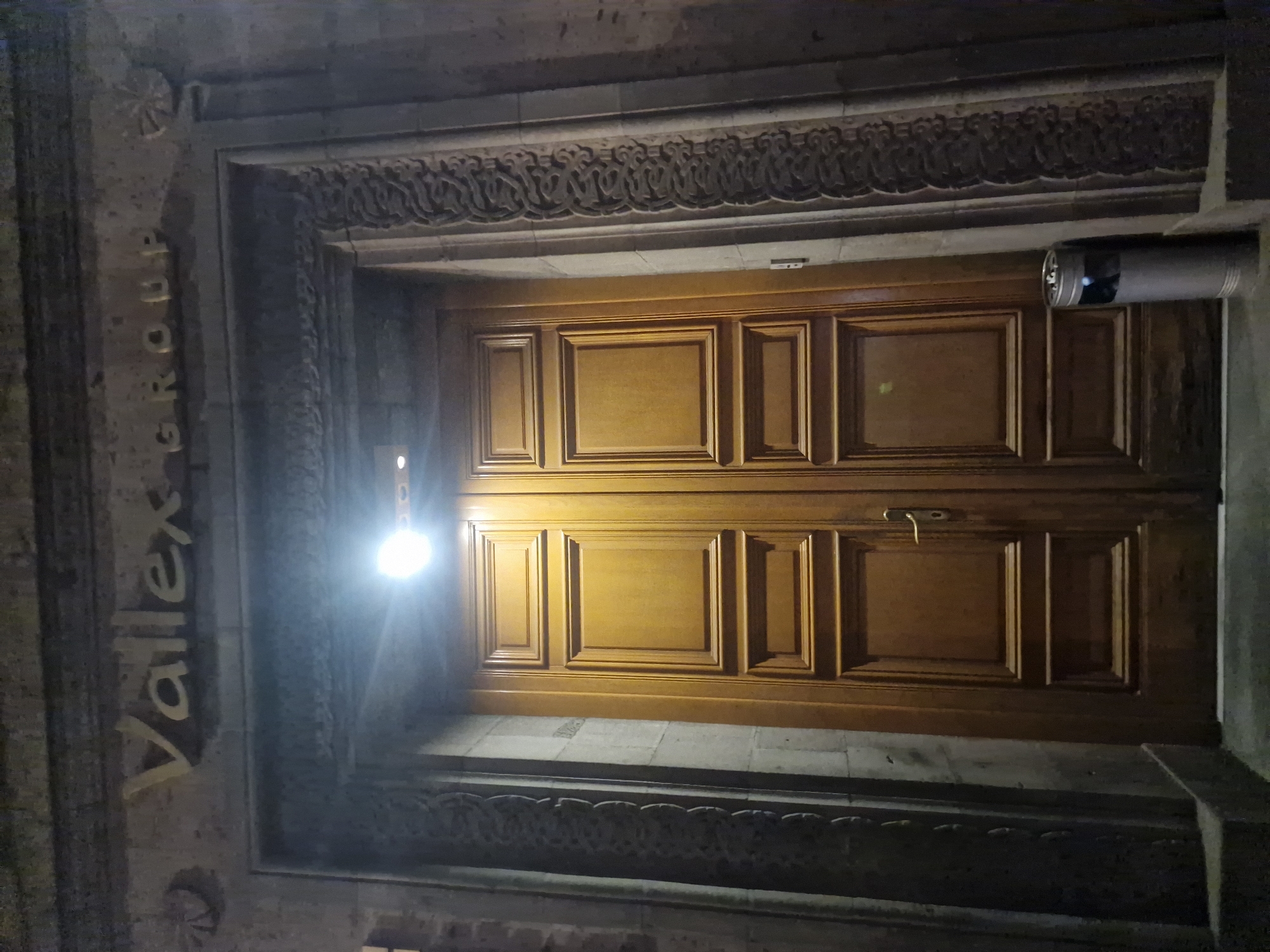 A wide-angle, low-light, eye-level shot captures a pair of imposing wooden double doors set within a stone entryway. Above the doors, on a stone lintel, is signage with the text "Vallex GROUP" in raised, gold-colored letters. The entryway is flanked by ornately carved stone pilasters, with decorative elements at the top corners of the lintel. A bright light source, possibly a spotlight, illuminates the area directly above the doors, casting a glare. To the right of the doors, at the base of the entryway, a silver-colored trash receptacle with a metal mesh top is visible. The overall impression is one of formal architecture and a nighttime setting.
