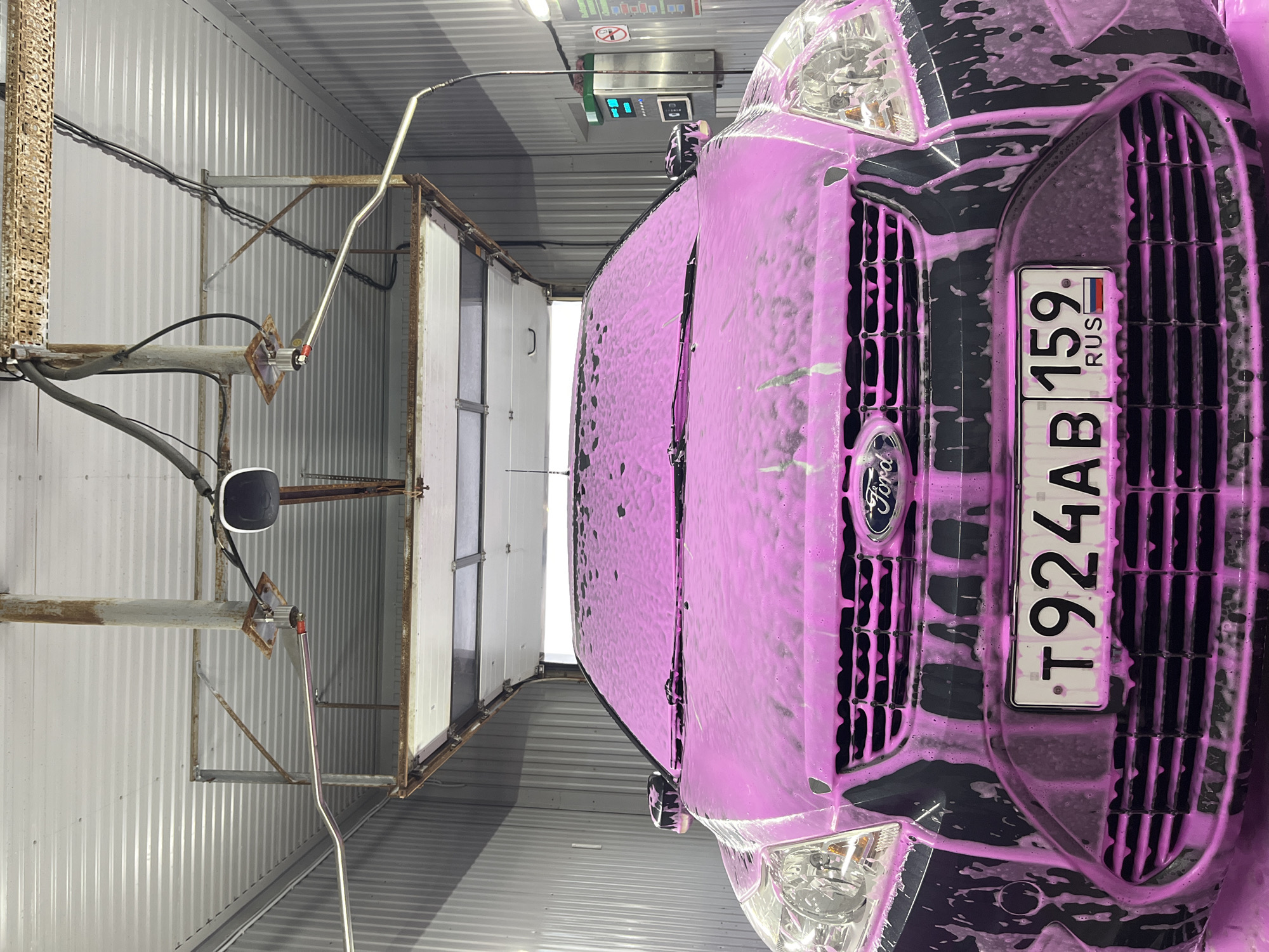 The image shows a black Ford van in a car wash. The vehicle is covered in pink soap suds. The license plate reads "T924AB 159 RUS". The car wash is located in Chaikovsky, Russia. The scene includes overhead cleaning equipment with nozzles and a pay station. The environment appears industrial, with metal corrugated walls and ceiling. There are no people visible.