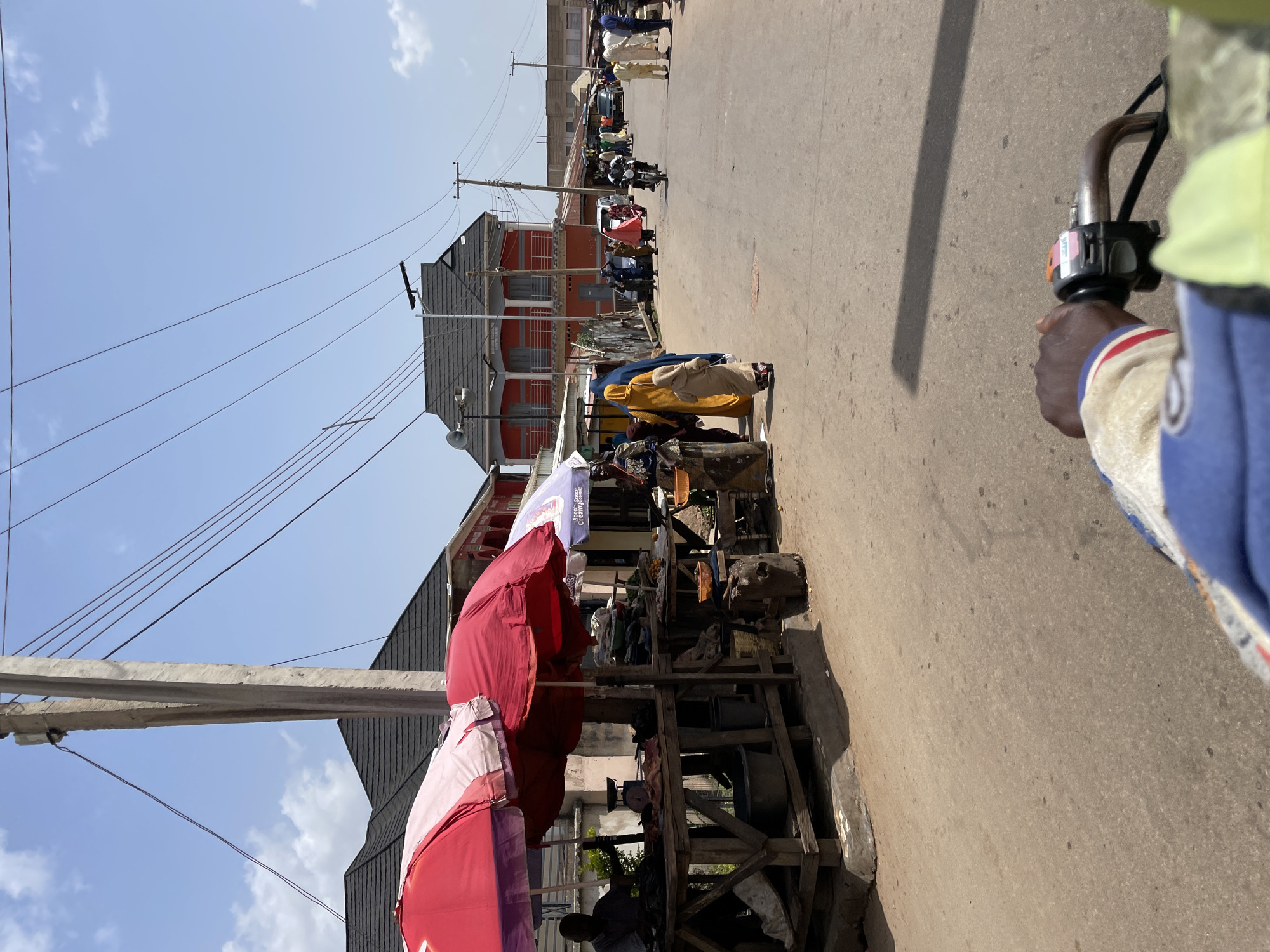 The image depicts a street scene in Gura Top, Nigeria. A person is holding the handlebar of a motorized vehicle, likely a motorcycle or tricycle, on the right side of the frame. The street runs parallel to the camera, with pedestrians and other motorized vehicles visible in the distance. Along the left side of the street, vendors have set up stalls under red and pink umbrellas, selling goods. Buildings are seen behind the vendor stalls, including a two-story structure with an orange facade. Utility poles with wires extend across the sky, which is light blue with some clouds.