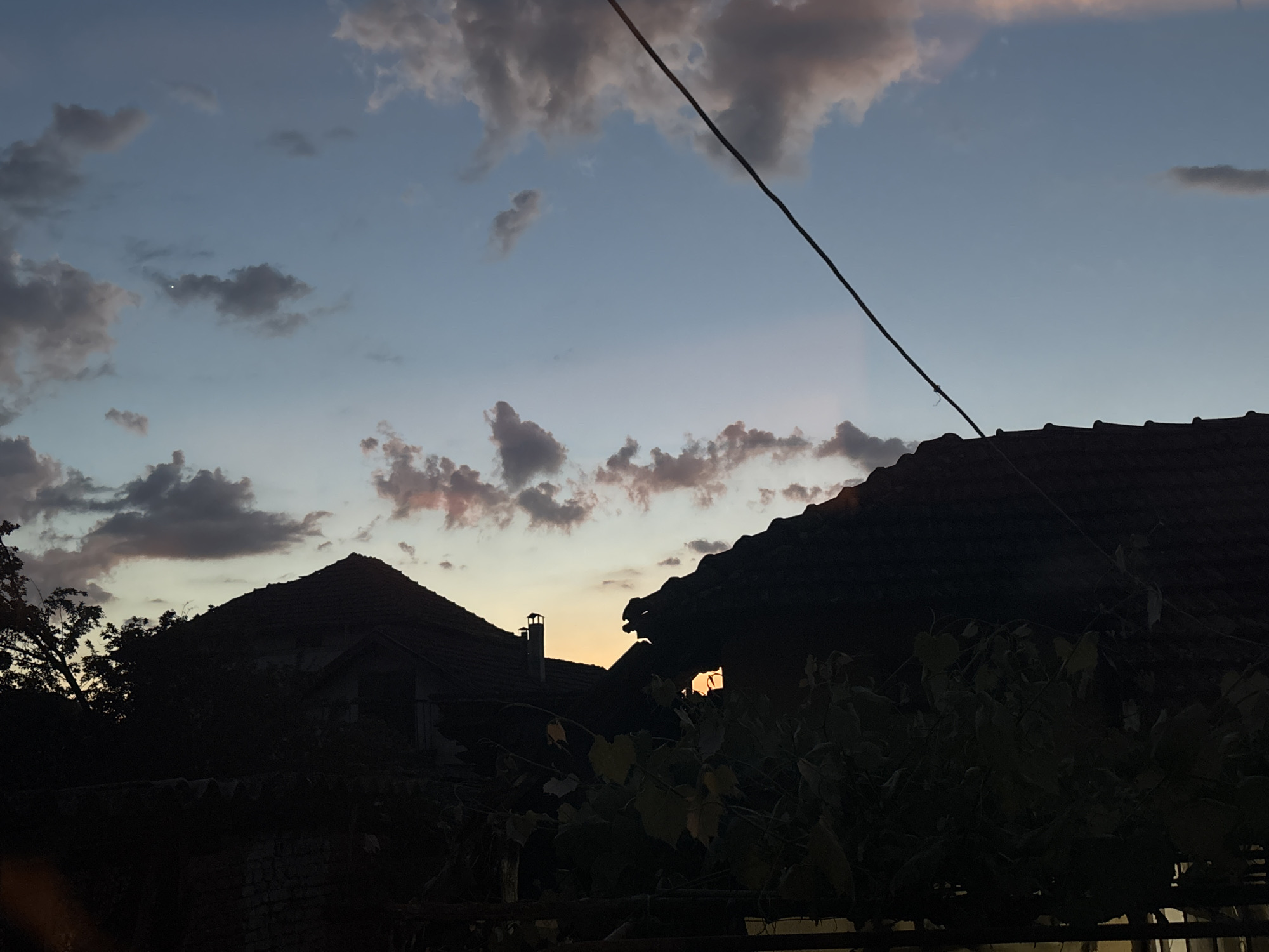 The image depicts a dusky sky with scattered clouds above the silhouettes of houses in an unknown location. The sky transitions from light blue to orange near the horizon. Overlapping rooftops with a chimney in between can be seen. A thick electrical wire stretches diagonally across the upper right quadrant of the scene. Some vegetation is visible at the bottom of the image.