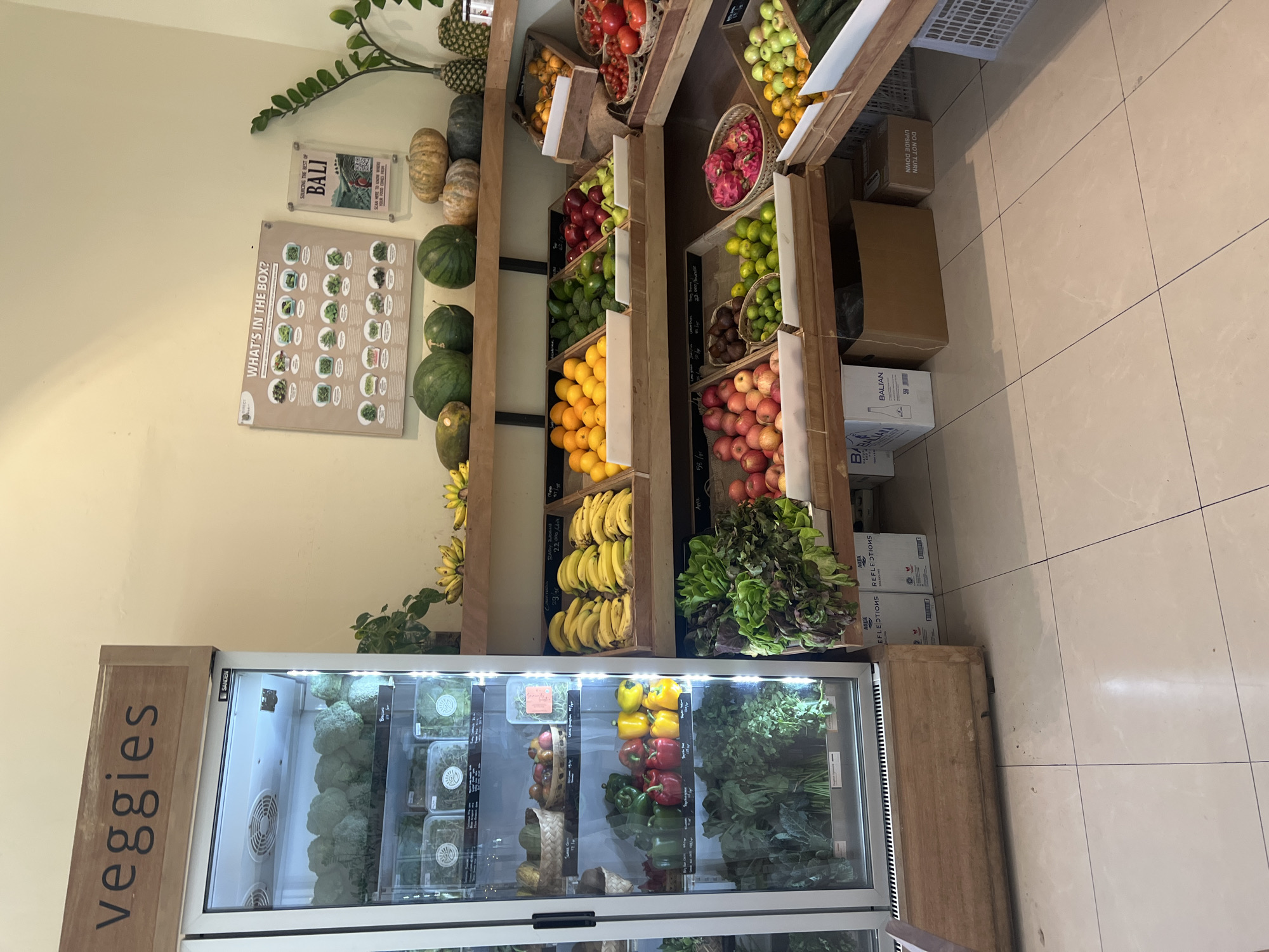 The image depicts the interior of a produce store in North Kuta, Indonesia. The store features a large refrigerated display case on the left, labeled "veggies," filled with various green vegetables, likely broccoli and leafy greens. Adjacent to the refrigerator are wooden shelves displaying an assortment of fresh produce. On the top shelf, bananas, watermelons, and pumpkins are visible. The middle shelves showcase fruits such as oranges, avocados, apples, and tomatoes, along with smaller fruits like limes and dragon fruit. The bottom shelves are stocked with more leafy greens, apples, and limes. Several cardboard boxes and plastic crates are placed on the floor beneath the shelves. The overall environment appears clean and organized, with bright lighting illuminating the produce.