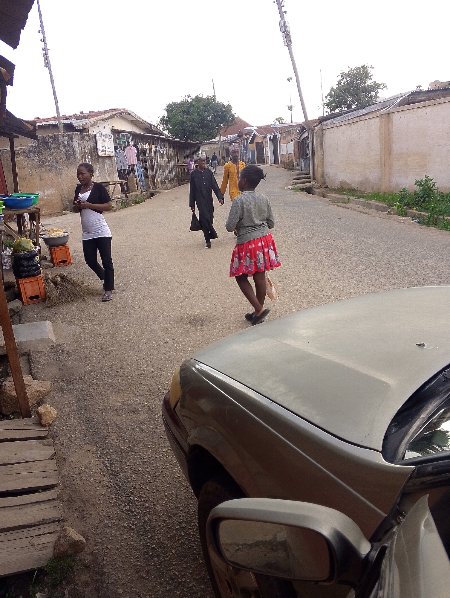 The image shows a street scene in Jos, Nigeria. The street is paved and leads into the distance, lined with buildings on both sides. A woman in a white top and black pants is walking along the left side of the road, passing a vendor selling food items from a makeshift stall. A man in a black robe walks toward the viewer with a bag. A child in a red floral skirt is also walking on the street, with another man in a yellow robe behind her. A light-colored car is parked on the right side of the frame, partially obstructing the view. Power lines are visible overhead. The buildings along the street are a mix of concrete structures and corrugated metal shacks.