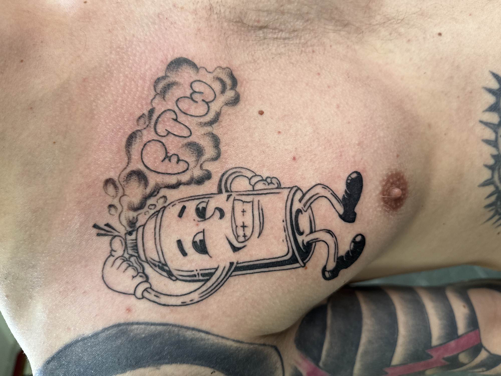 A close-up, chest-level shot captures the upper torso of a person, revealing a fresh, black and white tattoo. The skin around the tattoo exhibits some redness, consistent with recent tattooing, and shows fine hairs, pores, and several small moles. The person's right nipple is visible on the right side of the frame.
The main tattoo features an anthropomorphic character designed in a bold, cartoon style. The character resembles a cylindrical object, possibly a spray can or fire extinguisher, lying on its side. It has a cheerful, smiling face with wide eyes and a distinct nose. Its cartoon arms and legs are visible; the left hand holds a small object from which a cloud of smoke or steam billows upwards. Within this stylized smoke, the letters "CCP" are clearly formed. The character has a playful, relaxed posture.
A portion of another tattoo, featuring black and white stripes and a red lightning bolt, is visible on the person's right arm, partially entering the frame at the bottom. The image suggests an indoor setting. The location context is Moscow, Russia.