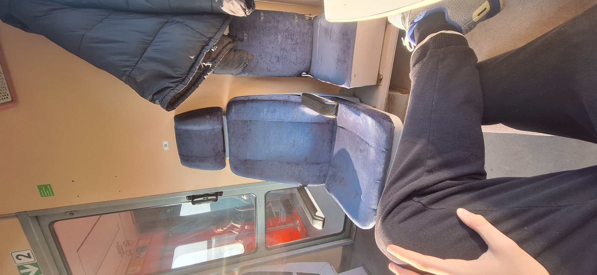 The scene appears to be inside a train car. There are blue upholstered seats, some facing each other, with a dark-colored jacket or coat draped over one set. A person is seated in one of the seats wearing black sweatpants and grey sneakers. A window is visible with sunlight streaming in. The walls of the train car are a pale orange. Small labels and signs are affixed to the wall near the window. Based on the layout of the train seats, this could be a regional or commuter train car. It is not possible to determine the precise location in the city of Burgas or even if the photo was taken within Burgas.