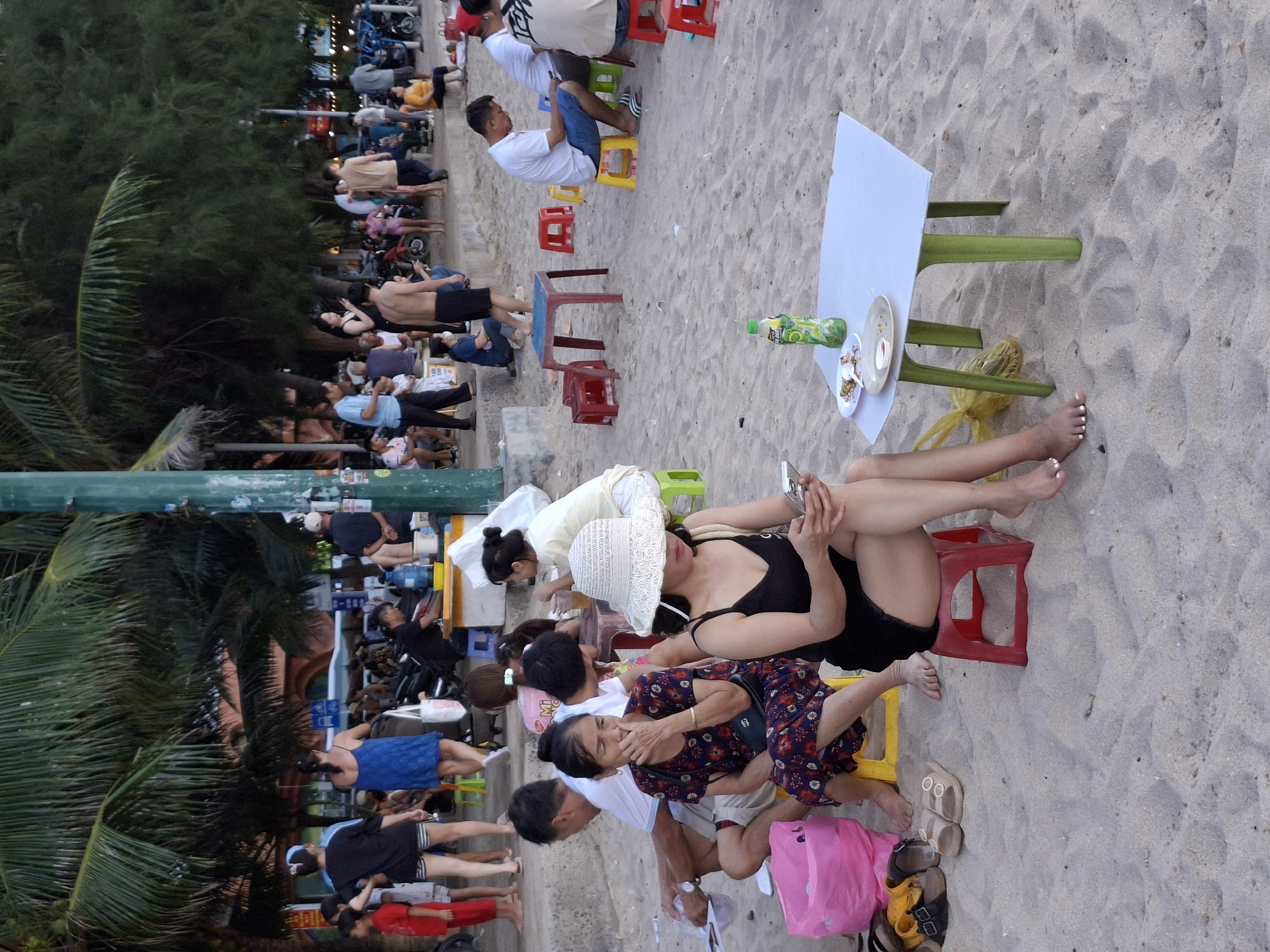 A group of people are gathered on a sandy beach, likely in Bac Nha Trang Ward, Vietnam, as indicated by the location context. The scene appears to be during the daytime, with overcast weather as suggested by the soft lighting and lack of harsh shadows. Some individuals are seated on low plastic stools and chairs around small tables, suggesting a casual gathering or a place to rest and observe.
In the foreground, a woman with a white sun hat is seated, wearing a black sleeveless top and black shorts. She is looking down at her phone. Next to her, another woman in a colorful dress is also seated, holding her hand to her face and looking forward. A plastic table with a light blue top and green legs stands nearby, holding plates with food and a bottle of green beverage.
Further back, more people are seen sitting and standing. Some are dressed casually in t-shirts and shorts, while others appear to be in swimwear. There are also several motorbikes parked along a walkway or road behind the seated groups, indicating that transportation is readily available. Palm trees and other lush green foliage are visible in the background, typical of a tropical beach setting. The overall atmosphere seems relaxed, with people enjoying a day at the beach.