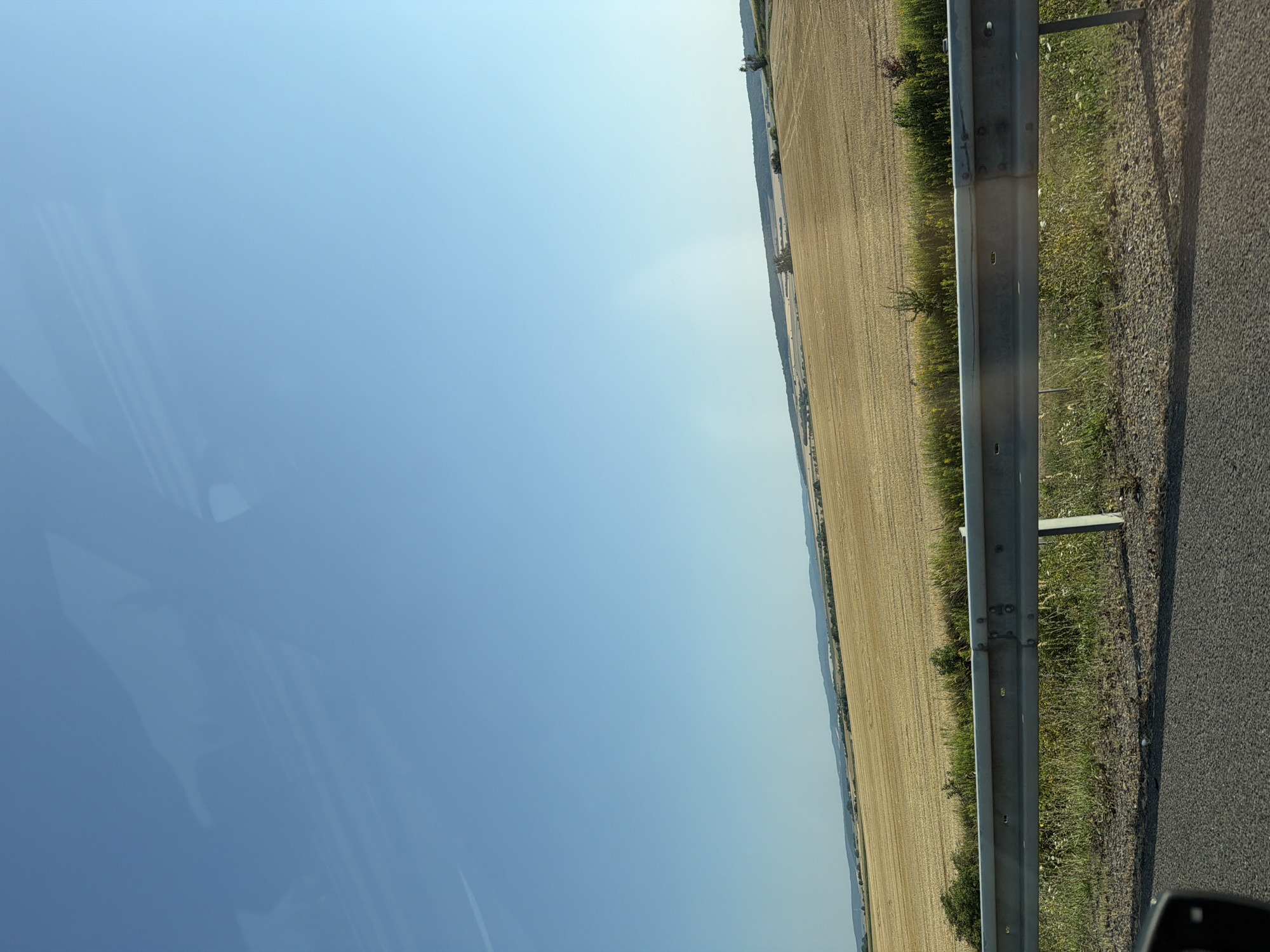 The image shows a roadside view of a harvested wheat field under a clear blue sky. In the foreground, a metal guardrail runs horizontally across the frame, separating the road from the field. The road surface is asphalt. The field is dry and golden, with rows indicating where the crops were harvested. In the background, a line of trees and hills can be seen. The scene is likely captured from a moving vehicle. The location is identified as Postoupky, Czechia.