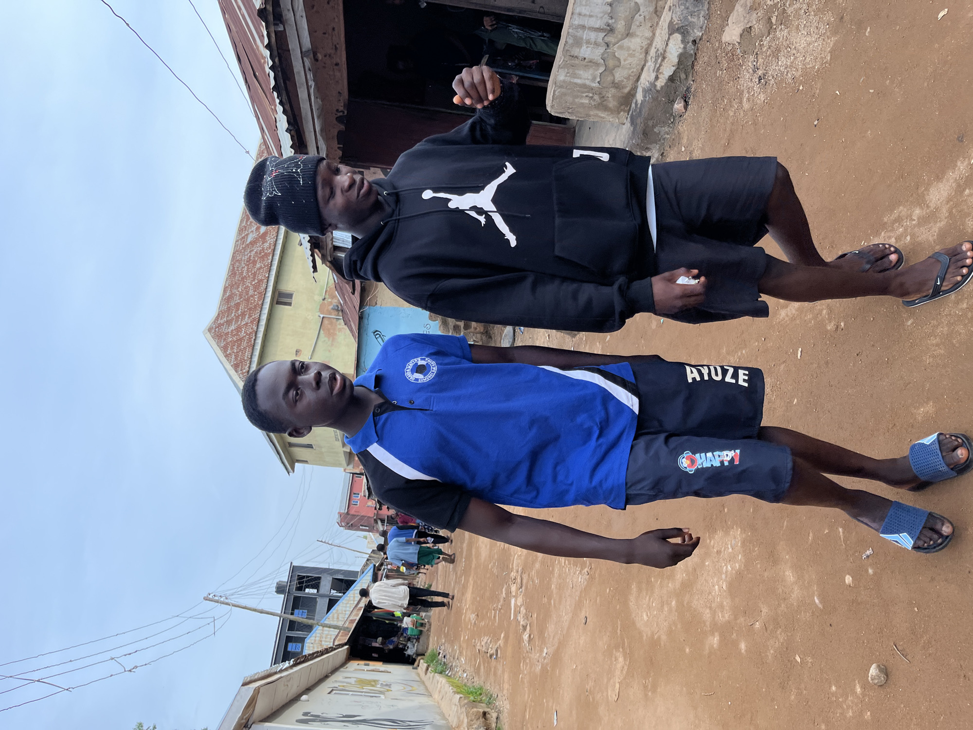 The image depicts two young Black males standing outdoors on a dirt path. The subject in the foreground is wearing a blue polo shirt with black and white accents and dark shorts. The subject behind him is wearing a black hoodie with a white Jumpman logo and black shorts. Both are wearing flip-flops.
The background suggests a bustling urban or semi-urban environment in Jos, Nigeria. Visible elements include corrugated metal roofing, simple buildings with light-colored walls, and power lines crisscrossing the sky. In the distance, there are more buildings and a glimpse of other people. The overall scene appears casual, with the individuals posing for the photograph. The lighting suggests it is daytime, likely overcast given the muted colors and lack of strong shadows.
There is text visible on the clothing of the subject in the foreground. On his shorts, the word "HAPPY" is clearly legible. The word "AYUZE" is visible on the waistband of the shorts. The subject in the background's hoodie has a prominent white Nike Jumpman logo.