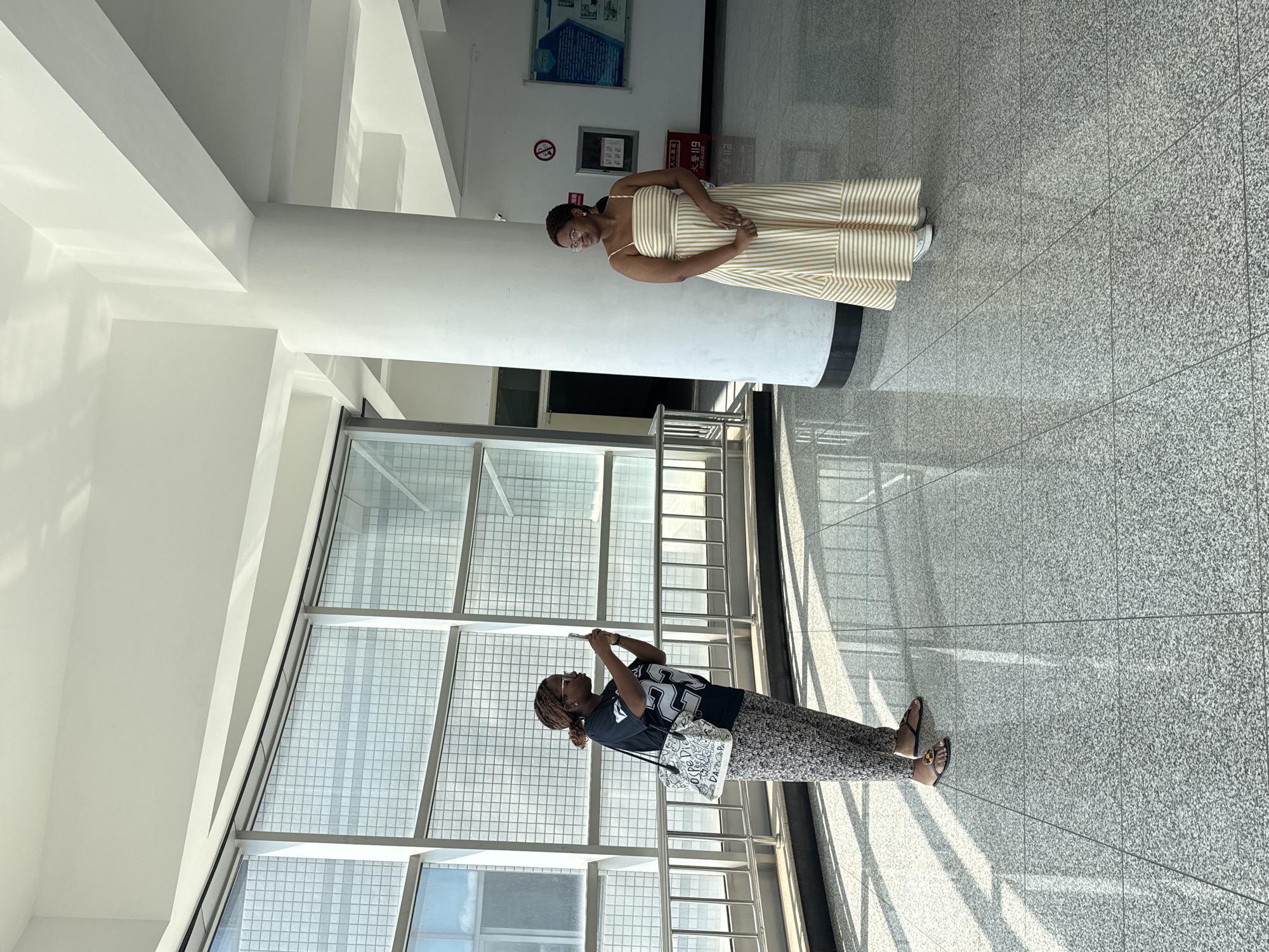 In the city of Guta, China, two Black women are in a brightly lit, modern indoor space with large windows and a tiled floor. On the left, a woman wearing a black t-shirt with a large white number "29" on it, floral patterned pants, and sandals is holding up a smartphone to take a picture of the other woman. She is also carrying a large tote bag with text and graphics. On the right, a woman in a sleeveless, floor-length striped dress with thin straps is posing and looking to her left. She is standing next to a large white pillar. The background shows large windows with a grid pattern, more pillars, and a fire alarm box on the wall. Sunlight streams in through the windows, casting shadows on the floor.