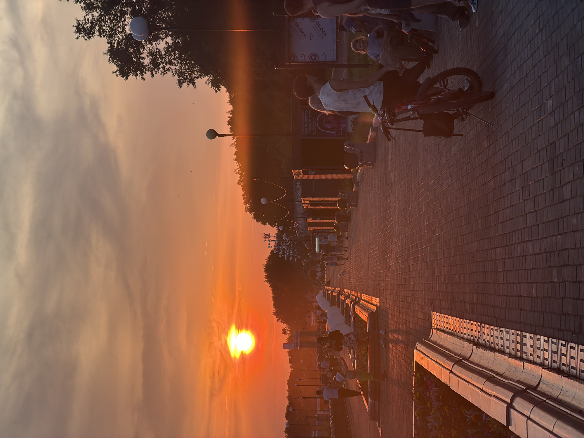 The image depicts a sunset over a paved promenade, likely in Birobidzhan, Russia. Several people are present, some walking, others sitting on benches, and one person is on a bicycle. The scene is bathed in the warm, golden light of the setting sun, casting long shadows and creating a serene, peaceful atmosphere. The sky is filled with soft clouds, tinged orange and pink by the sun.
There are lampposts along the promenade, and some trees are visible in the background. The ground is paved with brick-like tiles. On a sign in the background, the word "DILDEX" is visible twice, followed by a Russian word that appears to be "РЕАЛЬНОСТЬ". The overall impression is of a pleasant evening stroll or a moment of relaxation by the water or a park. The low angle of the sun indicates it is late afternoon or early evening.