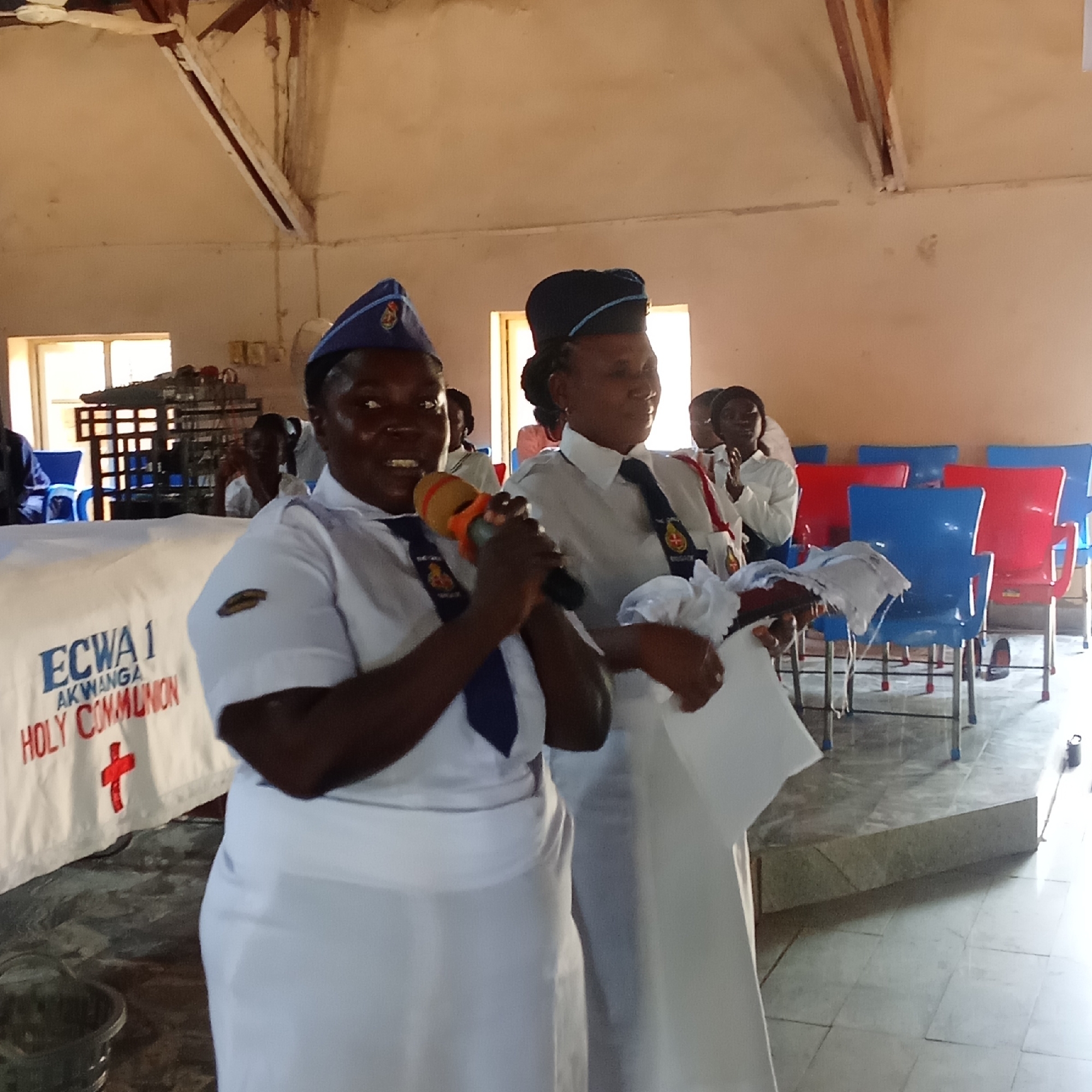 The image shows two women in white uniforms and dark blue hats standing in front of a table covered with a white cloth. The cloth has "ECWA 1 AKWANGA HOLY COMMUNION" printed on it, along with a red cross. The woman on the left is holding a microphone and speaking. The woman on the right is holding papers. The scene takes place indoors, likely in a church hall. Red and blue chairs are visible in the background. The location is Akwanga.
