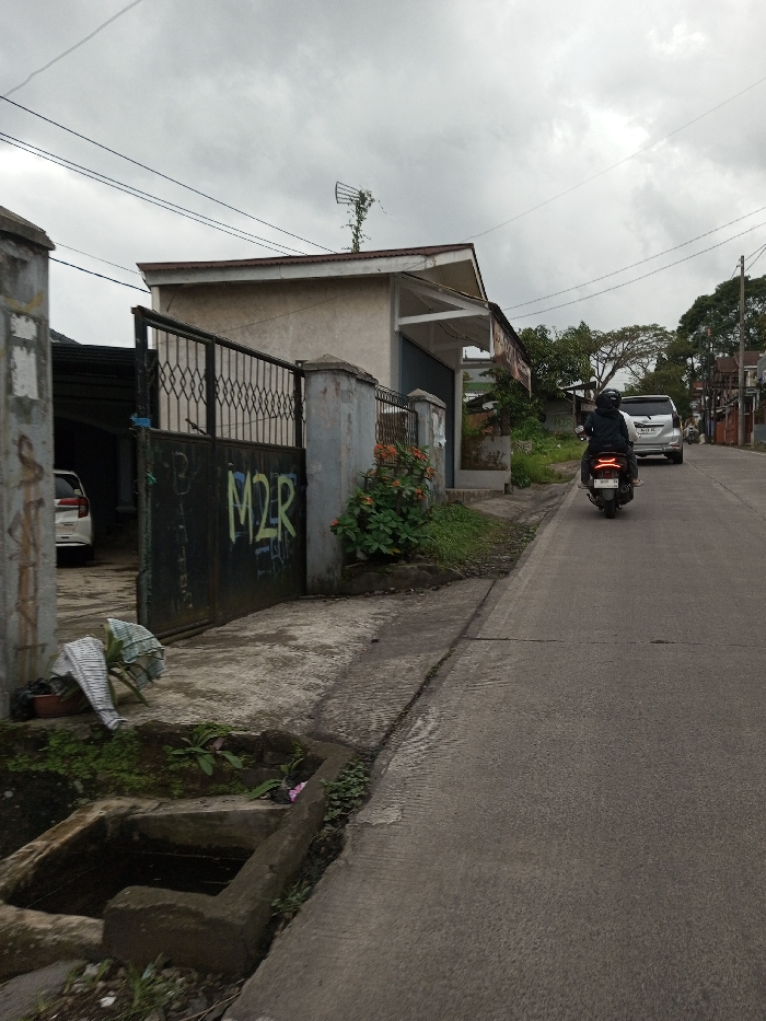 This image depicts an outdoor street scene in Melakudikadu, India, under an overcast sky. In the foreground, a concrete sidewalk runs parallel to a paved asphalt road, with a rectangular concrete drainage ditch containing standing water positioned on the left side of the sidewalk. Adjacent to the ditch, a brown pot holds a plant and a piece of white and striped fabric.
To the left, a dark metal gate, featuring a diamond pattern and yellow-green graffiti spelling "M2R," stands partially open, revealing a white vehicle parked behind it. The gate is supported by concrete pillars. Directly behind the gate and further right is a light-colored single-story building with a sloped roof. An antenna, partially obscured by vine-like vegetation, is visible on the roof. The building's facade includes a dark roll-up door or shutter. Reddish-orange flowering plants grow along the base of the building's fence line.
On the right side of the image, the asphalt road ascends slightly. A person wearing a dark jacket and helmet is riding a dark-colored scooter away from the viewer. Following the scooter is a silver or light-colored SUV. Utility poles and overhead power lines are visible, extending across the upper portion of the frame. The background shows additional residential structures and trees along the ascending road.