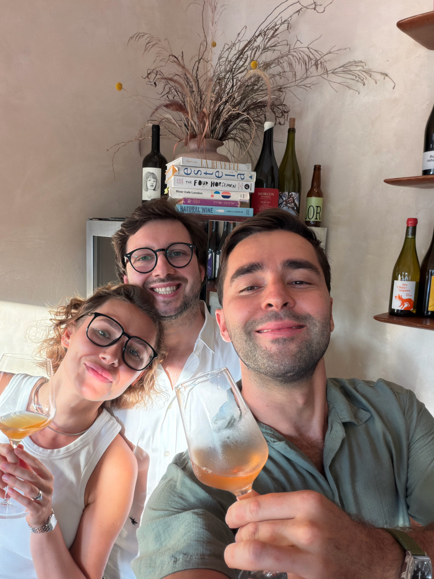 Three individuals, two men and one woman, are captured in a selfie-style photograph, holding wine glasses. The woman, on the left, wears glasses and a white tank top, with her curly hair framing her face. She is looking at the camera and smiling. To her right, a man, also wearing glasses, is smiling broadly. He has short dark hair and is wearing a white collared shirt. On the far right, another man is visible, looking directly at the camera with a slight smile. He has short dark hair and stubble and is wearing a green button-down shirt. All three appear to be holding glasses filled with a light-colored beverage, likely wine.
The background suggests an indoor setting with shelves displaying bottles of wine and stacks of books. A decorative arrangement of dried reeds and flowers sits on a shelf above the books. The wall behind them has a textured, light-colored finish. The lighting suggests it is daytime, with sunlight casting shadows. Based on the objects and style, this scene is likely located in a wine bar or a home with a curated display of beverages and reading material. The presence of books with Hebrew titles and the overall aesthetic are consistent with establishments found in Tel Aviv, Israel.