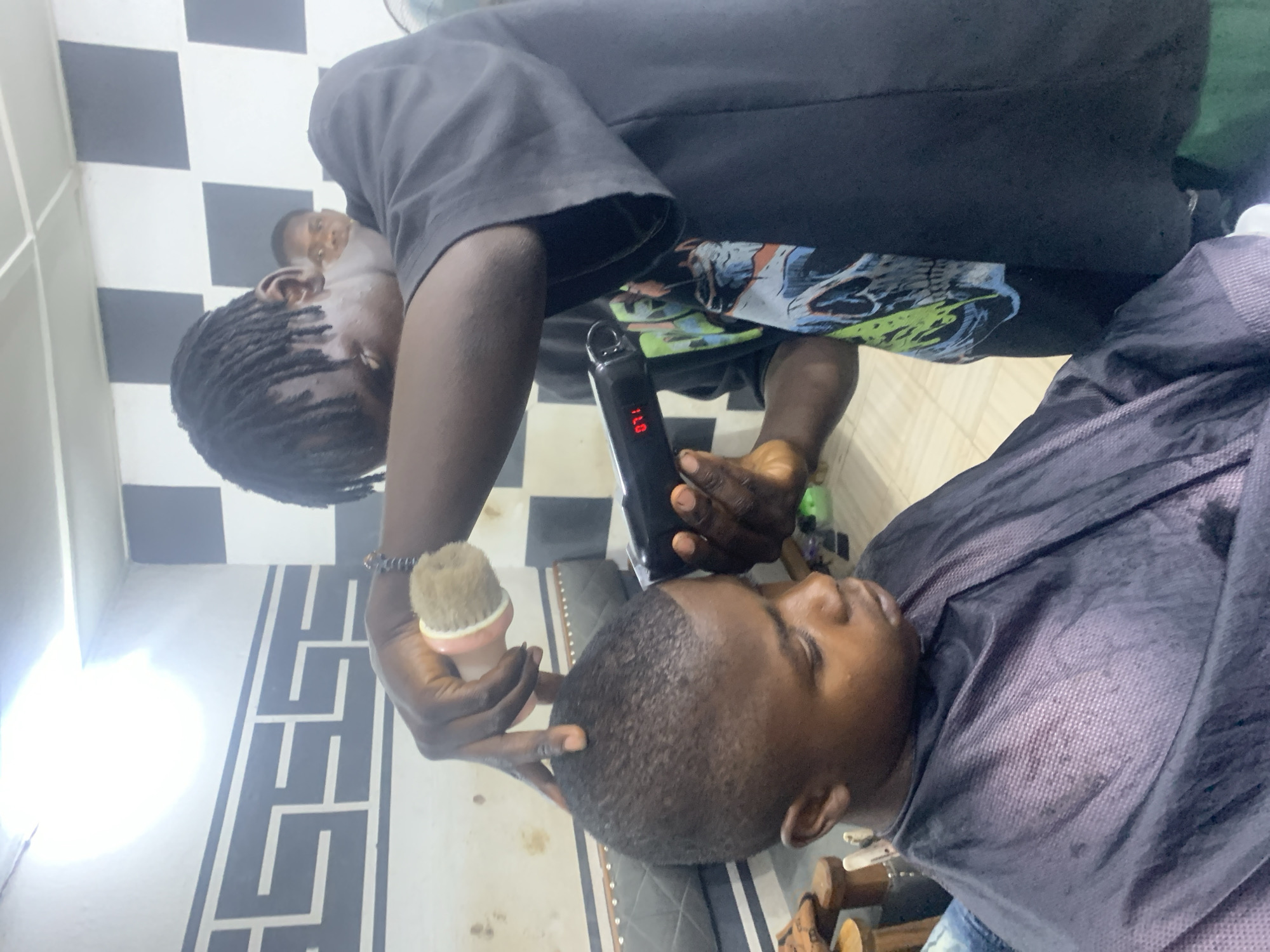 A barber with dreadlocks is giving a haircut to a client in a barber shop. The barber holds a hair clipper in his right hand and a brush in his left. The client is seated and covered with a black cape. The barber is wearing a black t-shirt and the wall has black and white patterns. The location is in Jos, Nigeria.