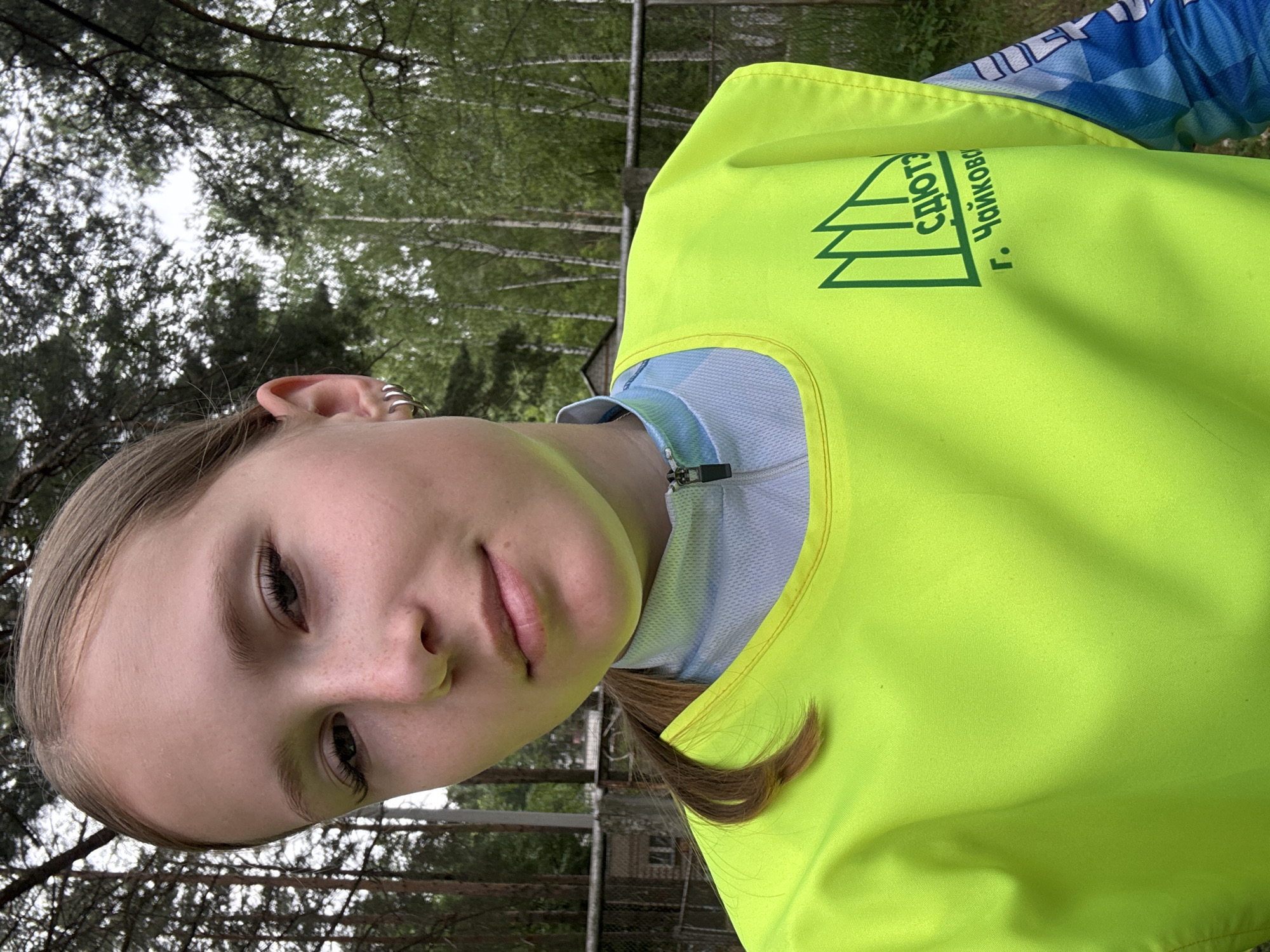 A young Caucasian female with blonde hair pulled back and light freckles across her nose is pictured in a medium shot, looking towards the camera with her head tilted slightly. She is wearing a bright neon yellow vest over a blue and white patterned athletic shirt with a zipped collar. The vest features a green emblem with text that reads "СДЮТЭ" and "г. Чайковский," indicating the location as Chaikovsky, Russia. The background consists of a blurred forest setting with tall trees and a fence. The overall lighting suggests an outdoor, daytime scene.