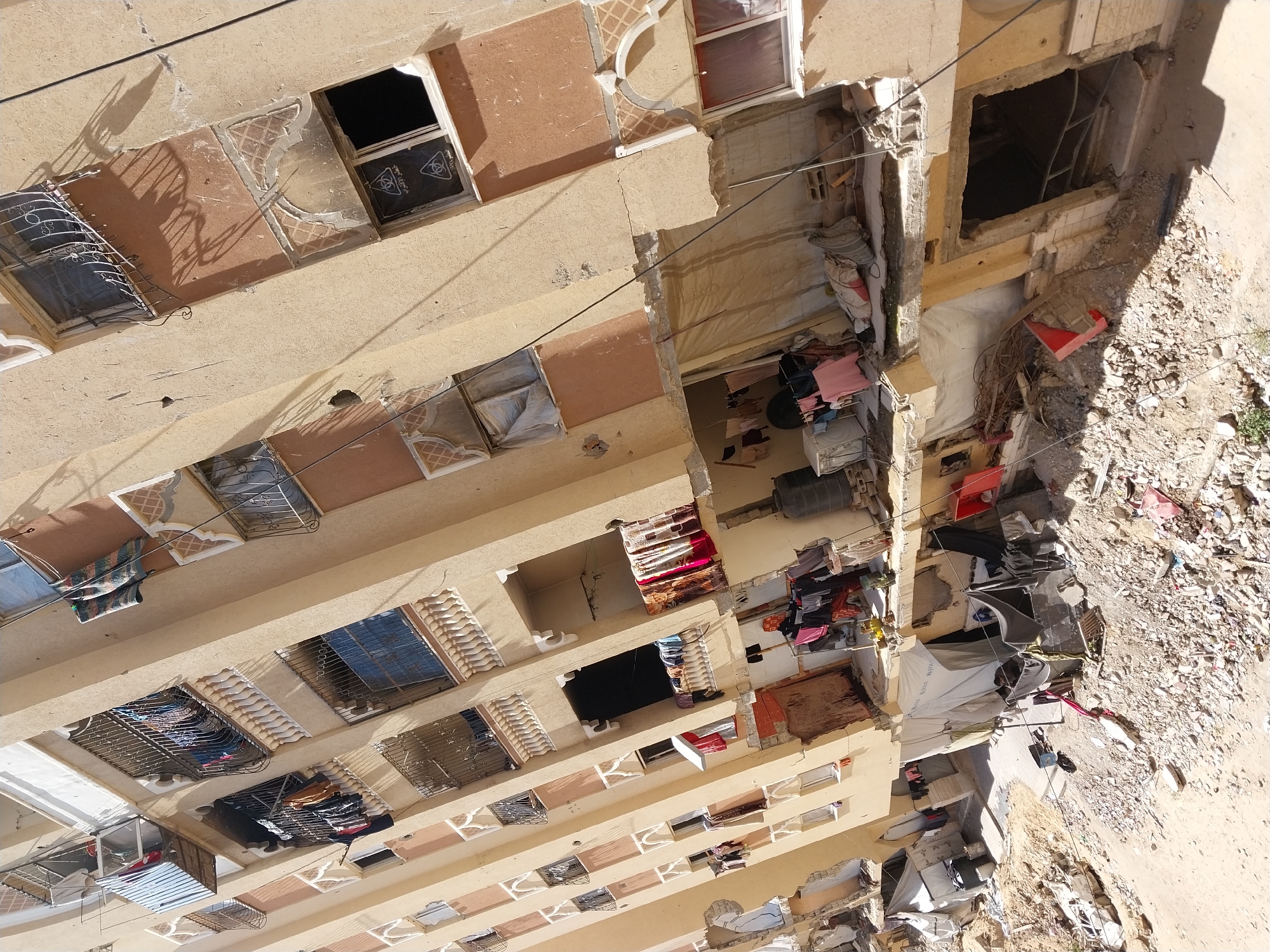 A high-angle shot depicts a building in Unknown, Unknown, revealing damage to its facade, exposing interior spaces. The building is a light tan color with multiple windows, some covered with protective bars or screens. Some window spaces appear damaged, with visible rubble and debris. Personal belongings such as clothing and household items are visible inside the damaged areas of the building. The ground adjacent to the damaged section is covered with debris, possibly from the building's collapse. No people are visible in the image.