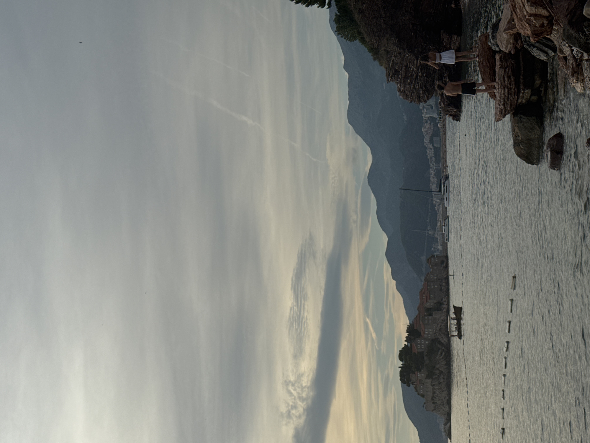 The image captures a coastal scene in Kamenovo, Montenegro. In the foreground, two individuals, a male and a female, stand on a rocky outcrop by the water. The male is shirtless, wearing dark shorts, while the female is wearing a white dress. The sea stretches out before them, with a small boat visible in the distance carrying several passengers. Across the water, a small island is adorned with buildings and trees. Beyond the island, a range of mountains forms the backdrop, with some distant buildings visible along the coastline. The sky is overcast with wispy clouds, and faint contrails are visible. The overall lighting suggests it might be late afternoon or early morning. The water appears calm, with a line of buoys marking a section of the sea.