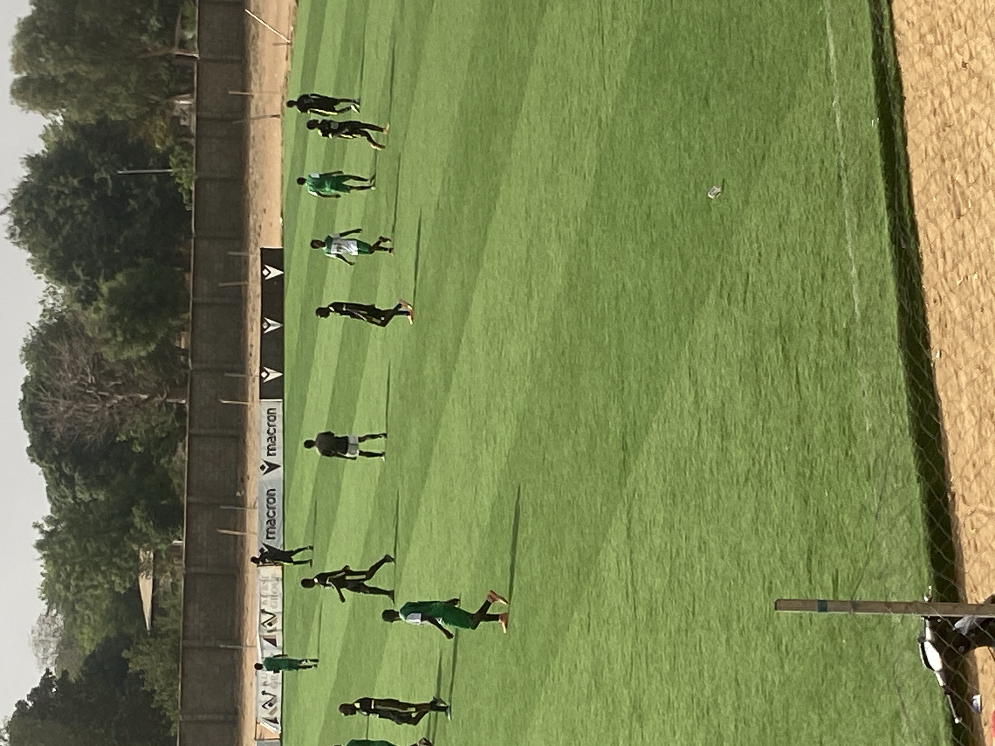 This image captures an outdoor football (soccer) pitch in Tudun Wada, Nigeria, under bright daylight. The main subject is a large, well-maintained green grass field, marked with faint white lines. Scattered across the pitch are approximately 15-20 individuals, predominantly male, dressed in football attire including jerseys and shorts. Some players wear dark-colored (black or dark green) kits, while others wear green or white jerseys, often with green accents. Their scattered positions and casual stances suggest they are engaged in training, warming up, or a light practice session, rather than an active match.
The bright sun casts long shadows from the players onto the grass, indicating a clear, sunny day, likely mid-morning or afternoon. Along the left side of the pitch (as oriented in an upright view), a low, light-colored wall or fence separates the field from a dense line of green trees and some dry, sandy ground, suggesting a rural or suburban setting. Advertising hoardings are visible along this edge, with the "macron" logo prominently displayed in white letters. On the far right, a section of a goal net and some light-colored netting are partially visible, along with an elevated, sandy area which could be part of a spectator stand. The overall scene depicts a functional football ground on a clear day.