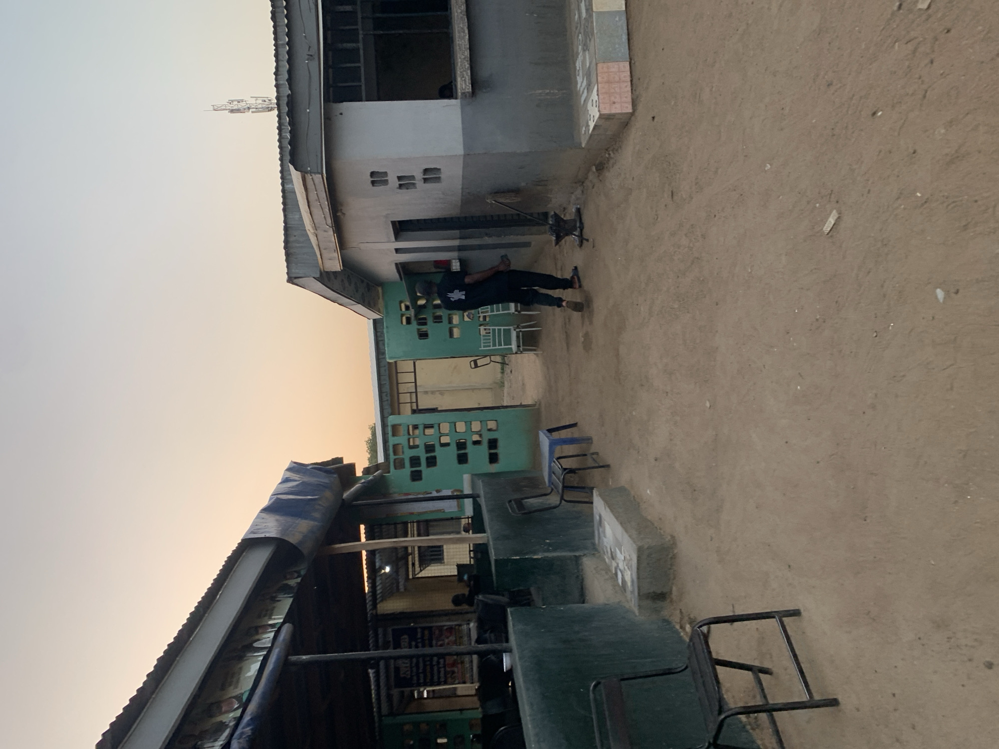 The scene appears to be an outdoor space in New Stadium, Nigeria. A man in dark clothing is walking or standing in the space between two buildings. One building is painted grey and white, with a visible window and multiple square openings. The other building, lower in height, has a blue tarp covering part of its roof and appears to be a commercial establishment, potentially a food stall or restaurant. Green tables and chairs are arranged outside this establishment. The ground is dirt or sand. A communication tower is visible on top of a building. The sky is light-colored, possibly indicating dawn or dusk.