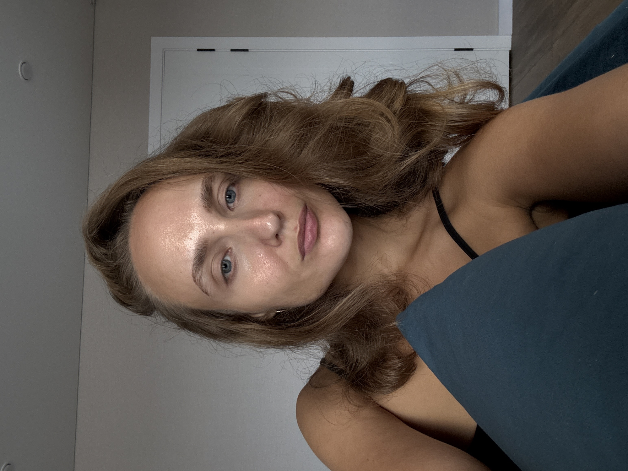 A woman with light brown hair and blue eyes is reclining on a teal pillow in what appears to be a room. She is wearing a black tank top. Her hair is styled in loose waves. The background shows a plain wall and a closed door. The lighting suggests an indoor setting, possibly during the day. There is no visible indication of the location being in the city of Подшивалово, Russia.